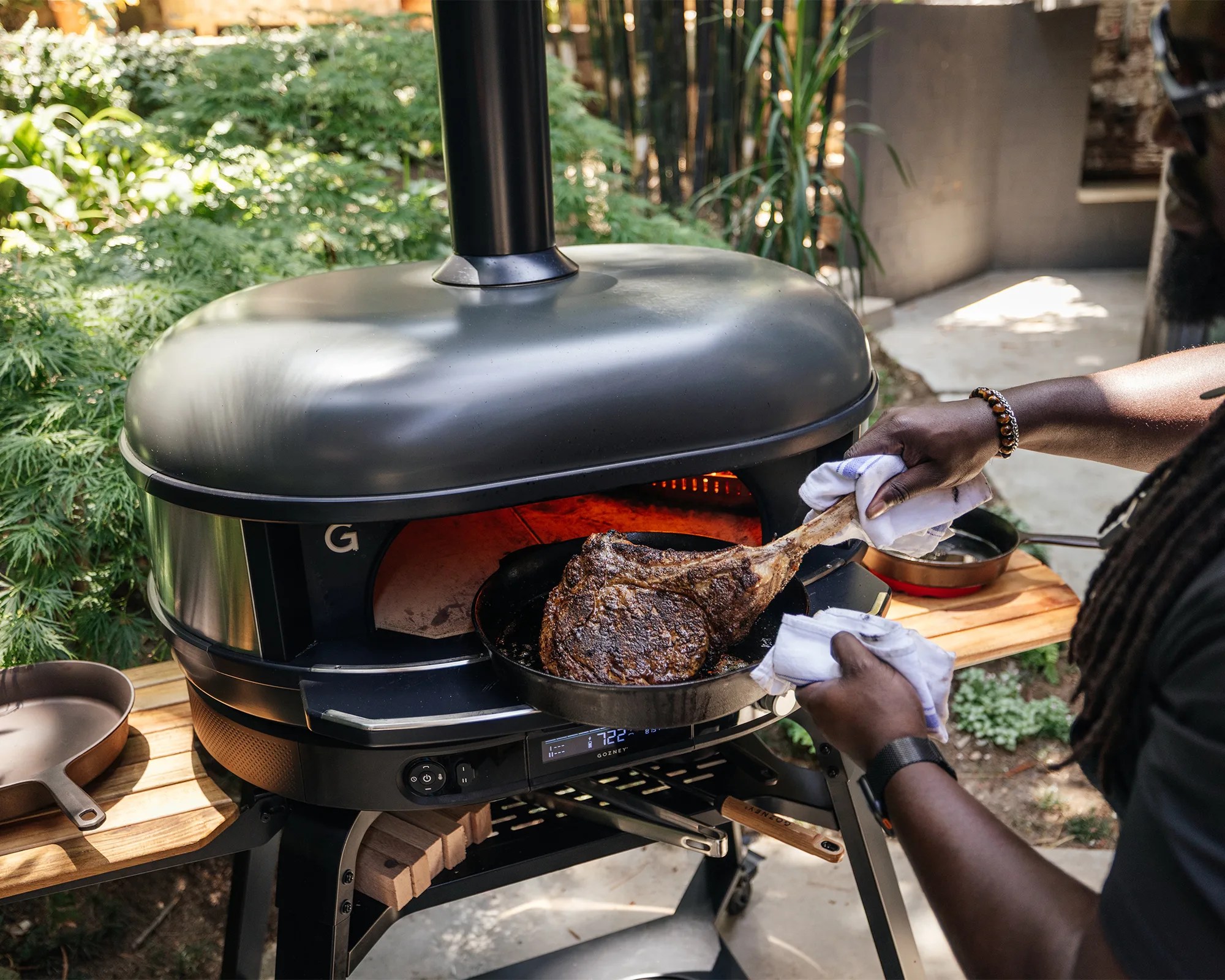 A person is holding a cast iron skillet with a large cooked tomahawk steak, using a cloth to handle the hot pan. The skillet is placed on the edge of a modern outdoor pizza oven or grill with a digital temperature display showing 722°F. The scene is set outdoors with greenery in the background.