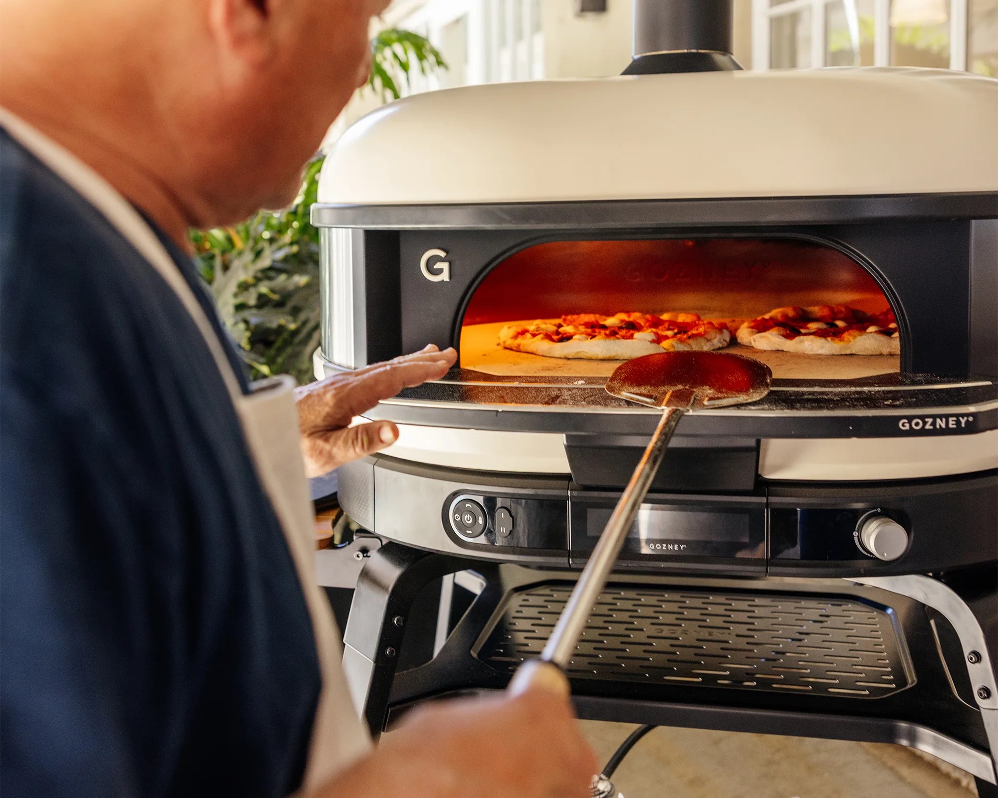 A person is using a pizza peel to place or remove two pizzas inside a Gozney outdoor pizza oven. The oven has a black and cream color scheme with a visible control panel and a chimney on top. The scene appears to be set in a bright, outdoor or semi-outdoor area.