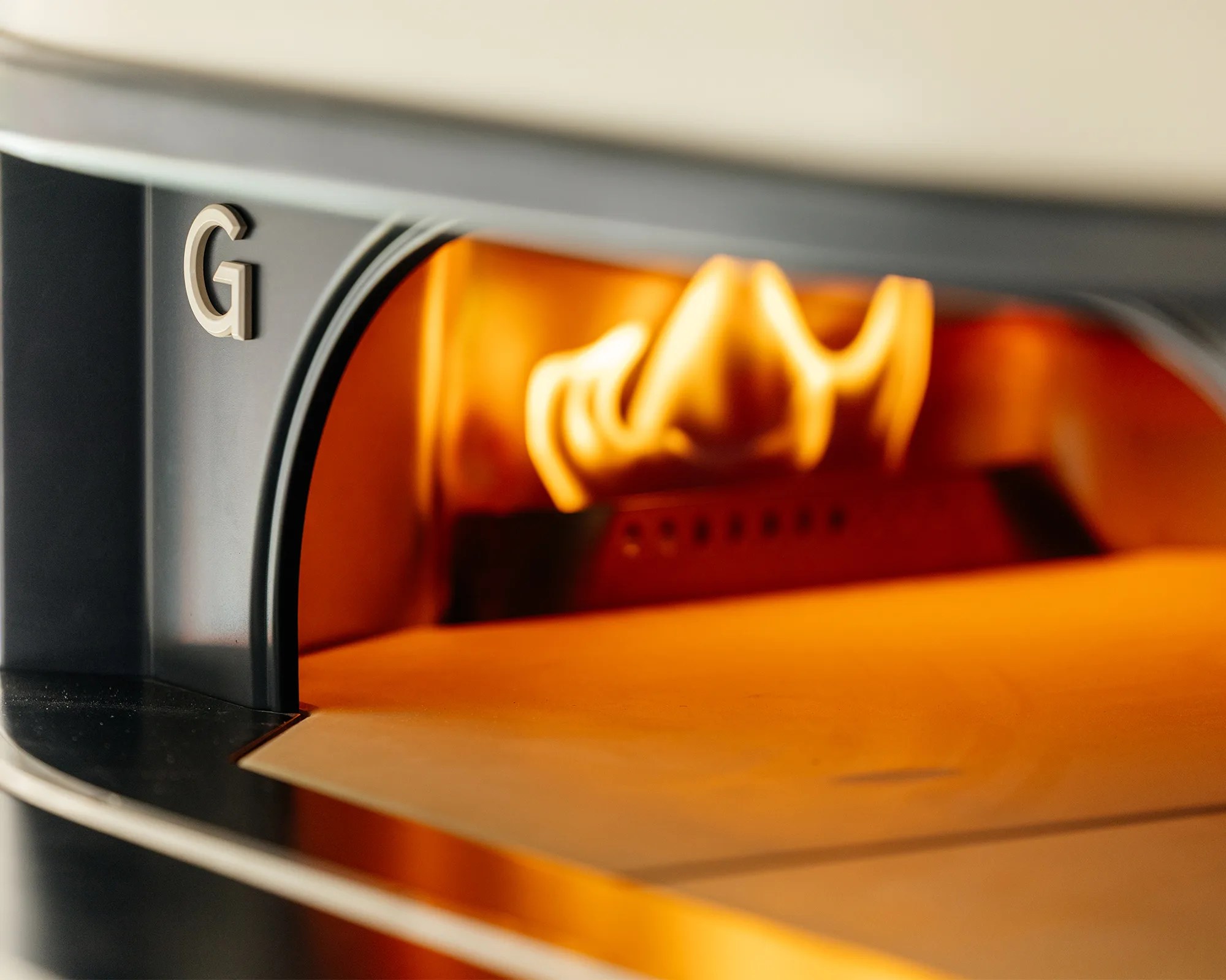 Close-up of a modern pizza oven with a visible flame inside, featuring a black exterior and a metallic letter "G" on the left side. The oven interior glows with warm orange light from the fire.