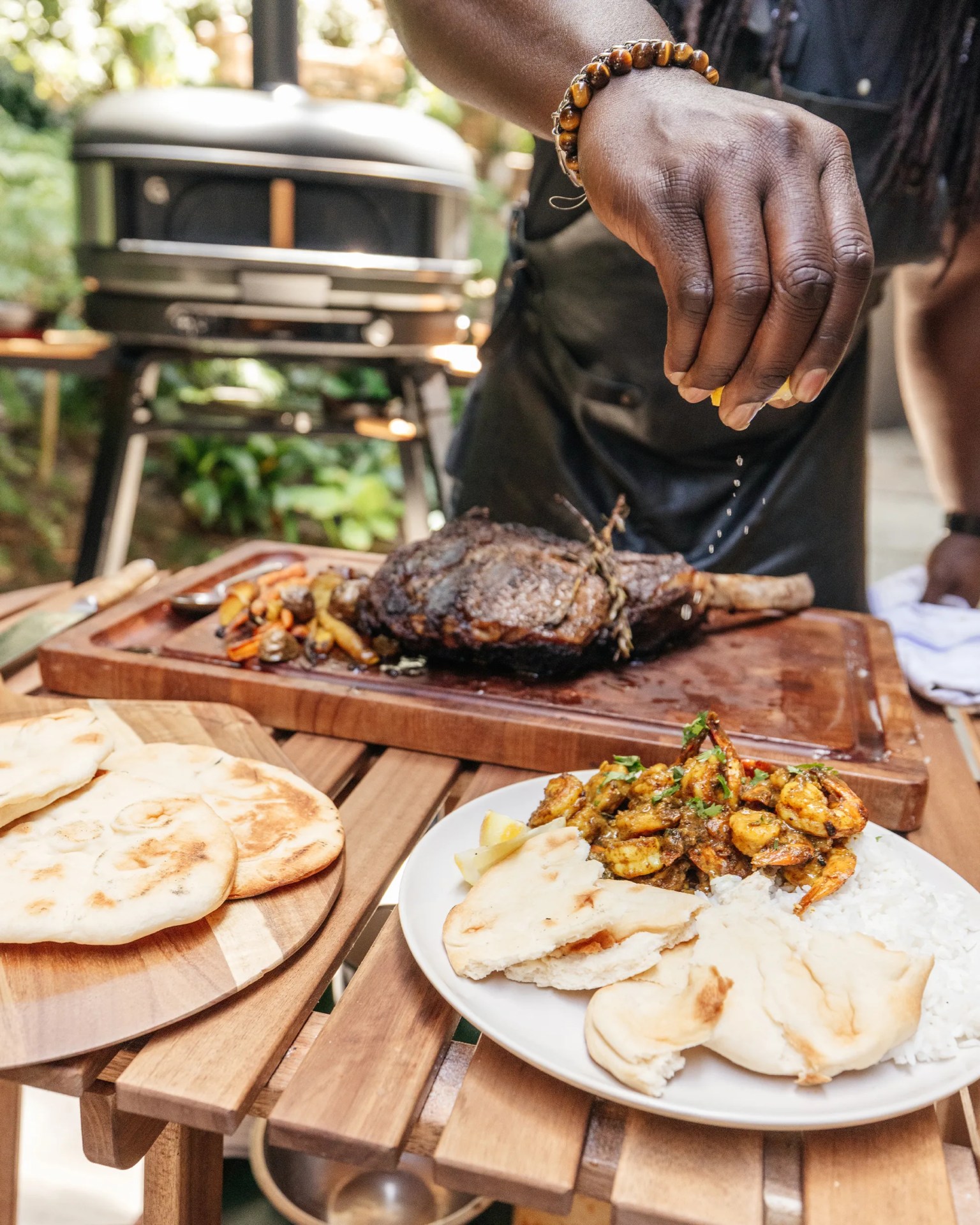A hand is squeezing lemon juice over a plate of cooked shrimp garnished with herbs, served with white rice and pieces of flatbread. In the background, there is a wooden cutting board with a large grilled steak and roasted vegetables, and a grill is visible further behind. Another wooden plate with flatbread is on the table. The setting appears to be outdoors.
