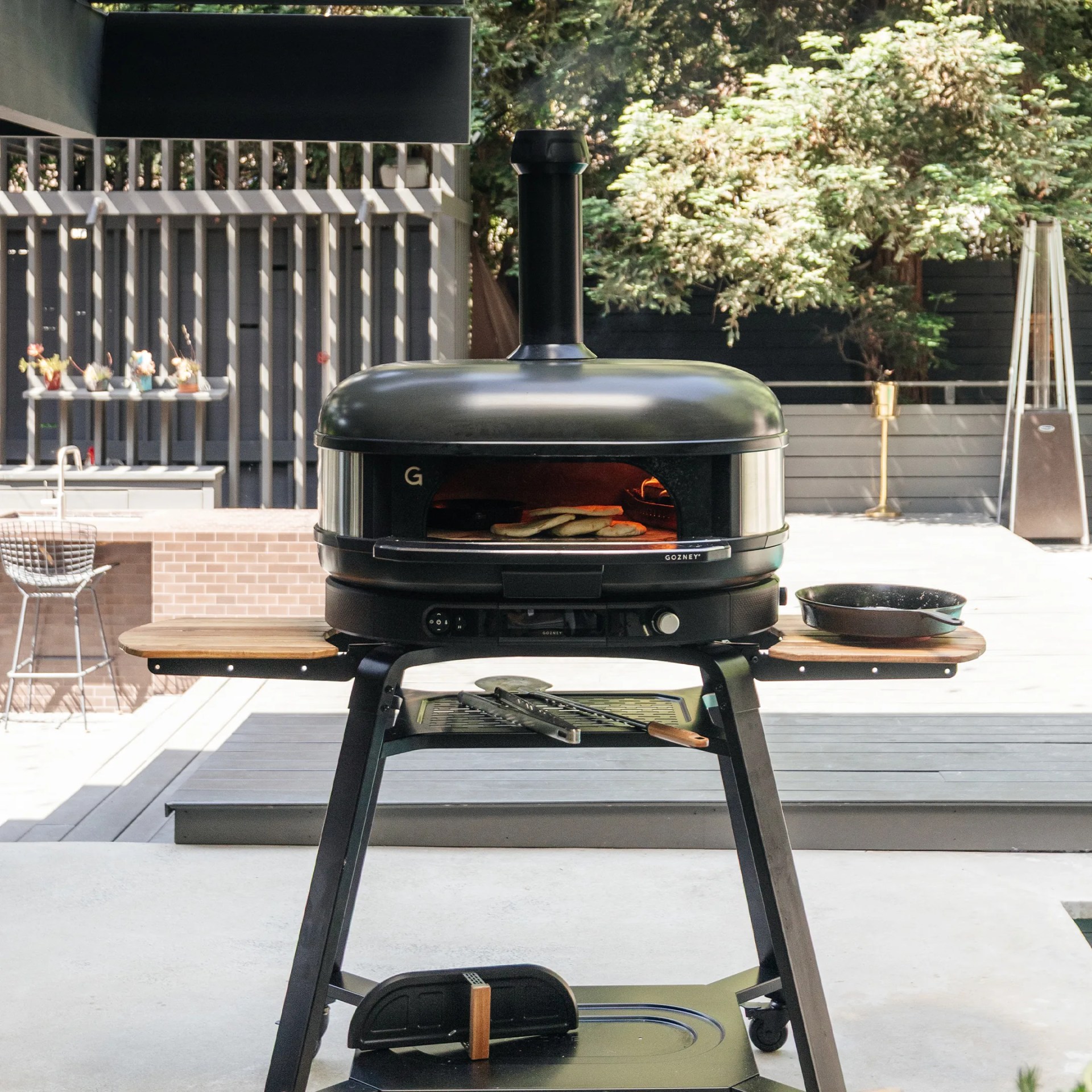 Outdoor black pizza oven with a chimney, standing on a metal frame with wooden side shelves. Inside the oven, several pizzas are baking. A cast iron skillet is placed on the right wooden shelf, and grilling tools are resting on a lower metal rack. The background shows a modern patio with plants and outdoor furniture.