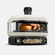 A modern, compact wood-fired pizza oven with a cream-colored dome and black base. The oven has a front opening showing flames inside, a digital temperature display on the left, a storage area with stacked firewood in the center, and a control knob on the right. The brand name "Gozney" is visible on the front.