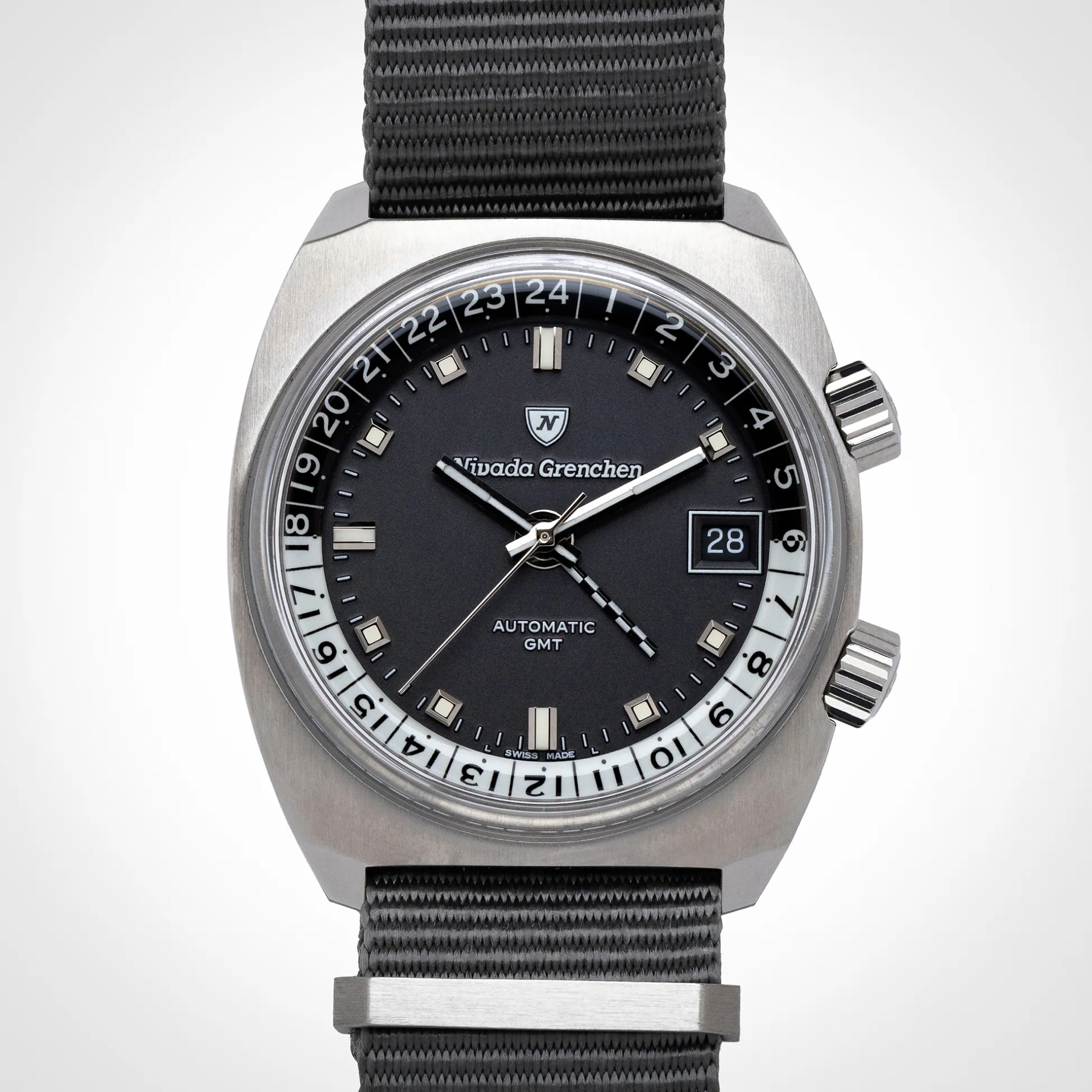 Nivada Grenchen automatic GMT wristwatch with a black dial, luminous square and rectangular hour markers, and a date window at 3 o'clock. The watch features a 24-hour rotating bezel with black and white sections, two crowns on the right side, and a gray fabric strap. The hour, minute, second, and GMT hands are visible, with the GMT hand having a checkered pattern. The case has a brushed stainless steel finish.