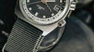 Close-up of a wristwatch with a black dial featuring silver square hour markers, a date window displaying "28," and the text "AUTOMATIC GMT" and "SWISS MADE." The watch has a brushed stainless steel case, two crowns on the right side, and a textured gray fabric strap.