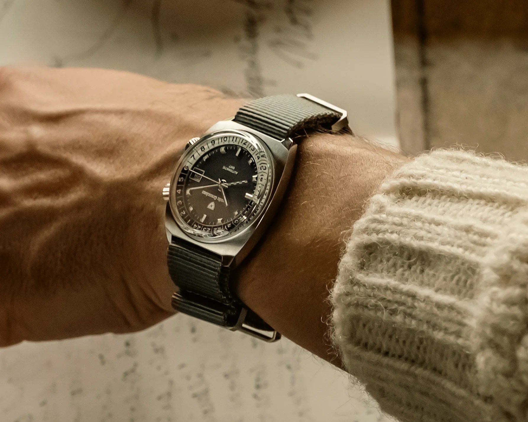 A wristwatch with a black dial and silver case is worn on a person's wrist. The watch has a gray fabric strap and features a date window at the 6 o'clock position. The person is wearing a cream-colored knitted sweater. The watch face includes a 24-hour scale on the inner bezel and has two crowns on the right side of the case.