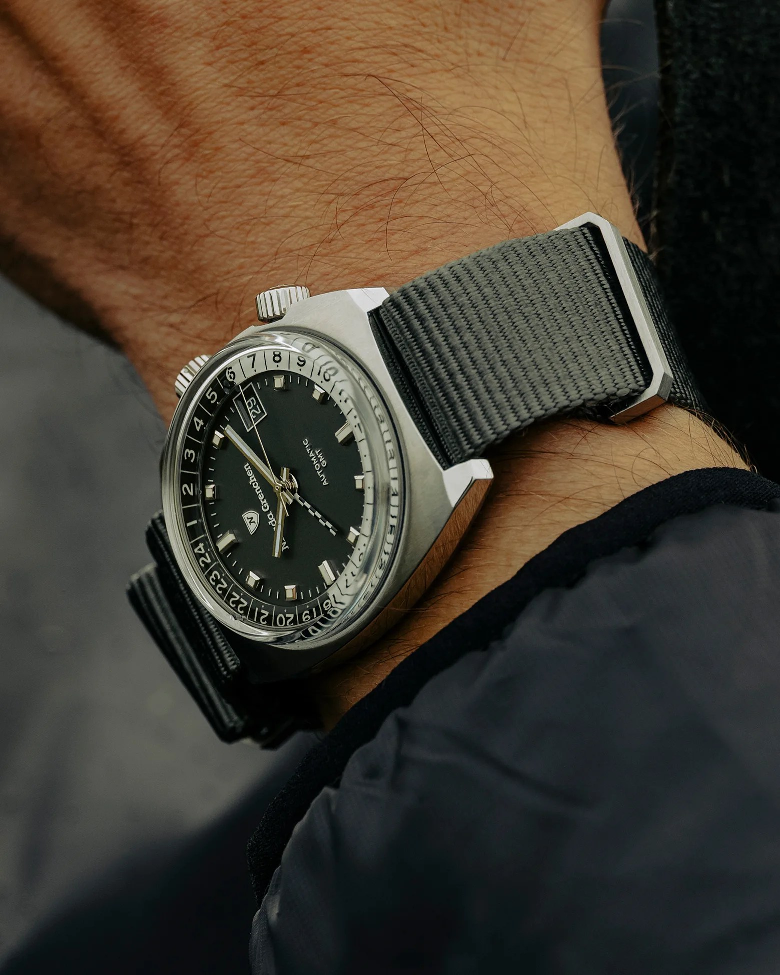 Stainless steel wristwatch with a black dial and silver hour markers, featuring a date window at the 3 o'clock position. The watch has two crowns and a rotating inner bezel with 24-hour markings. It is worn on a textured gray fabric strap. The watch face displays the brand name "Monta de Grenchon" and the words "Automatic GMT.