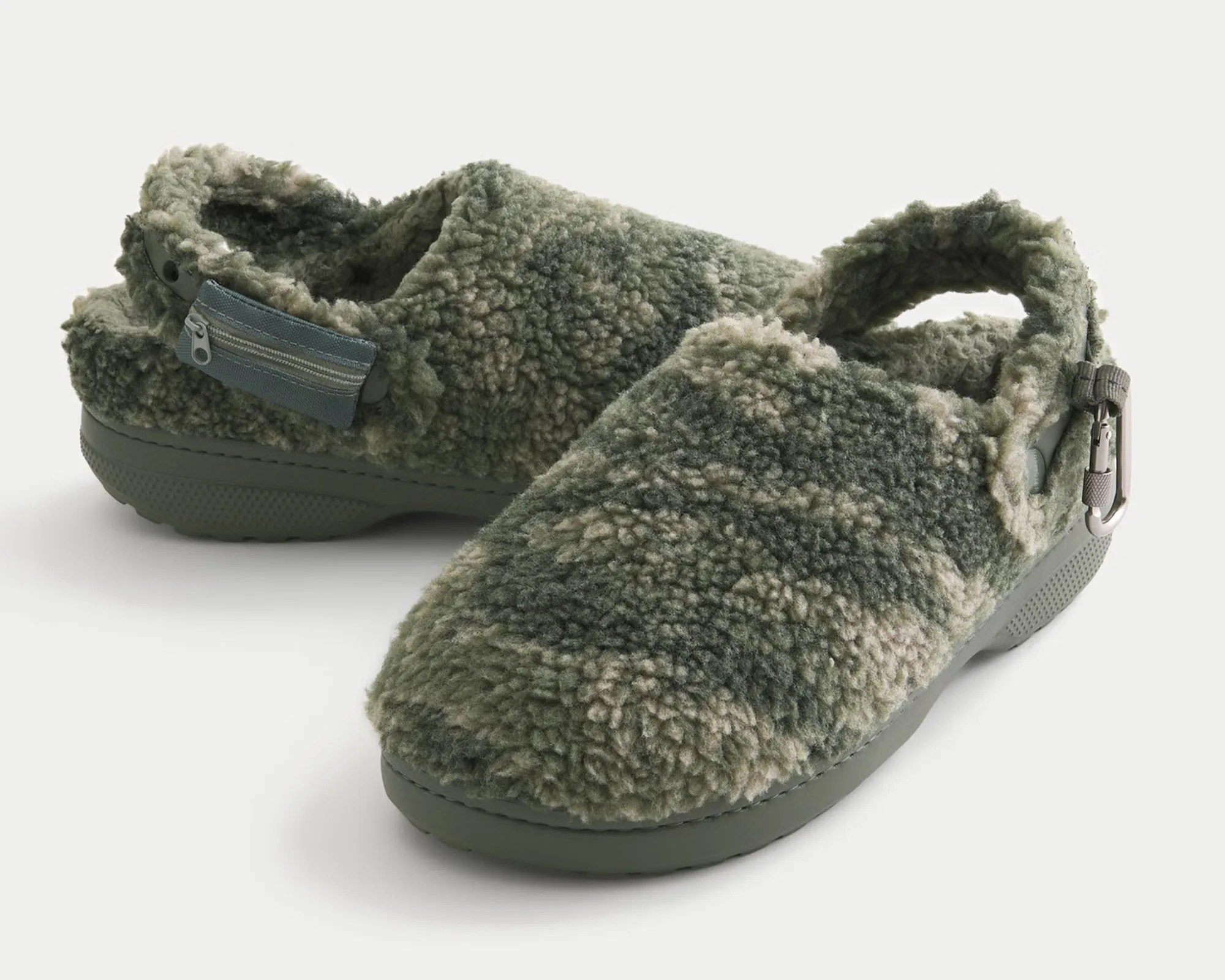 Pair of green and beige camouflage-patterned fuzzy slip-on clogs with a textured sole, featuring a small zippered pocket on the side of one shoe and a metal clip attached to the back strap of the other.