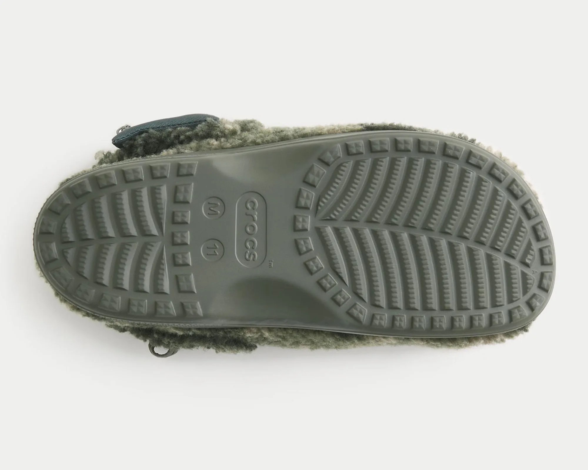Sole of a Crocs shoe with a textured tread pattern, featuring the Crocs logo and size markings "W 11" and "M 11." The shoe has a fuzzy, wool-like outer material in a greenish-gray color.