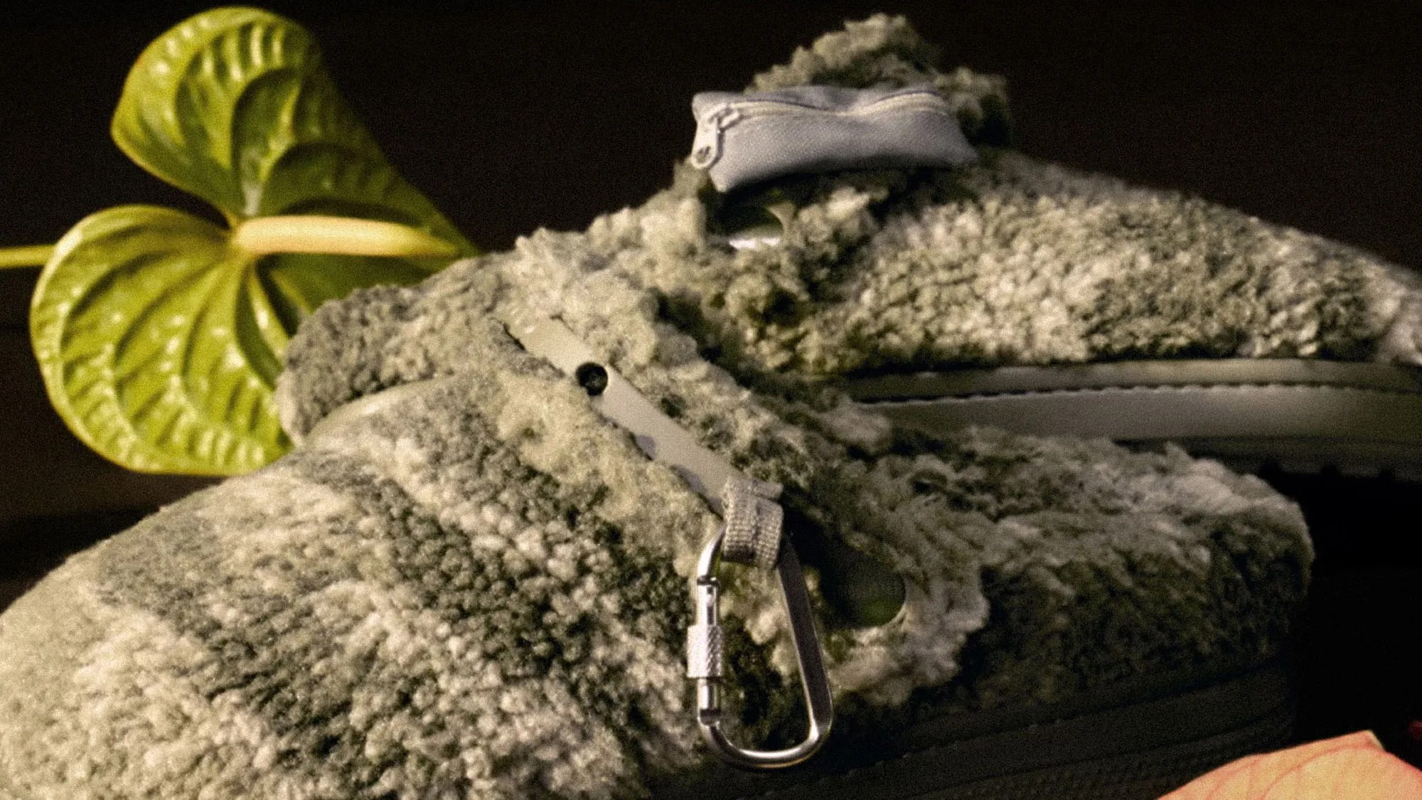Close-up of a pair of fuzzy, textured shoes with a gray and white wool-like material. The shoes feature a small metal carabiner attached to a strap on the side. A green leaf is visible in the background.