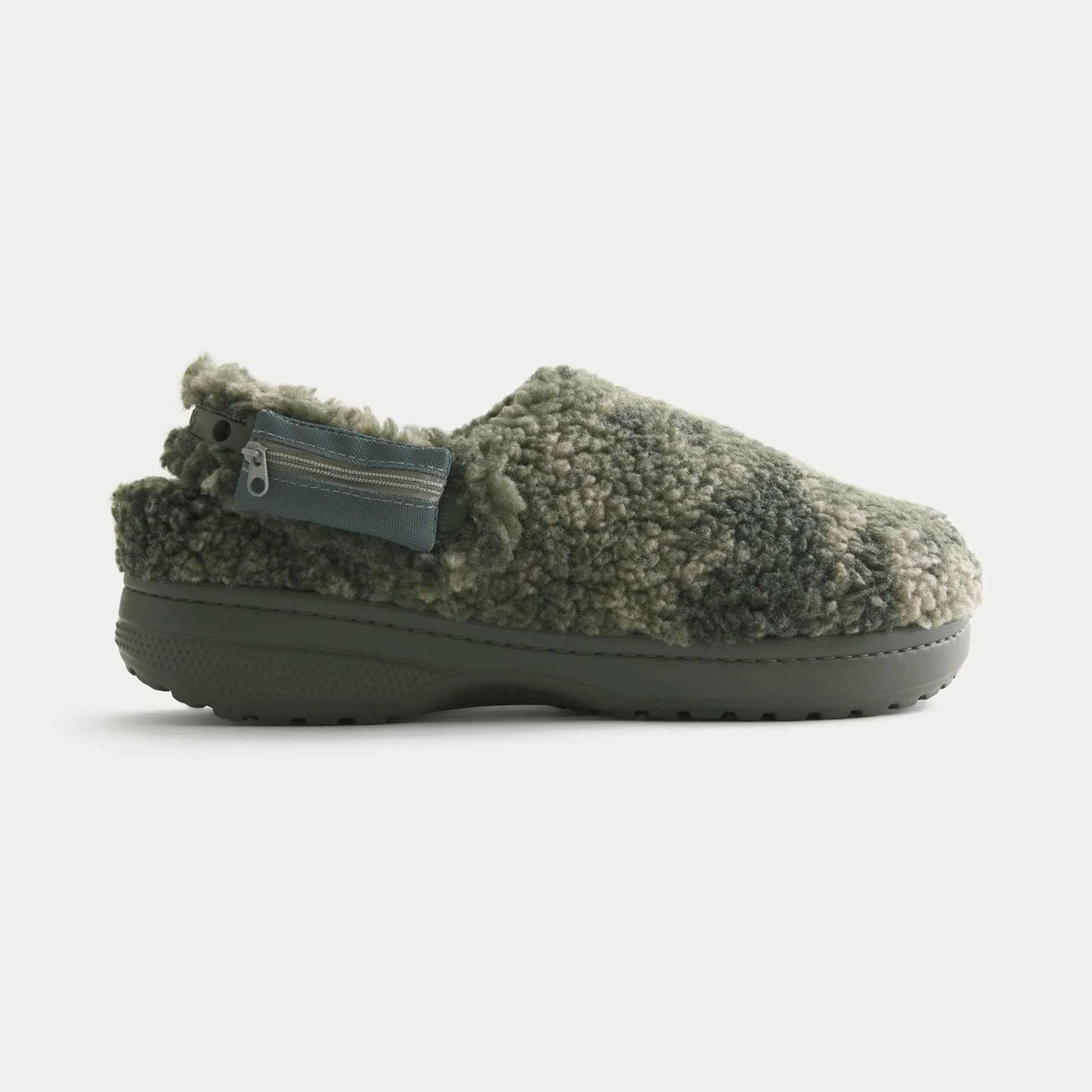 A slip-on shoe with a thick, textured, green and beige fleece upper. It has a solid green rubber sole and a small zippered pouch attached to the back strap. The shoe appears cozy and warm, designed for comfort.