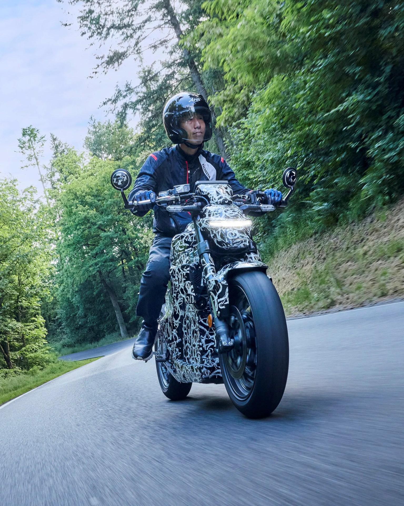 A person wearing a black helmet, blue gloves, and a dark jacket is riding a motorcycle with a black and white abstract camouflage pattern on a winding road surrounded by green trees. The motorcycle has a bright horizontal LED headlight and round side mirrors. The rider's feet are positioned on the footrests, and the scene appears to be in a forested area during daylight.