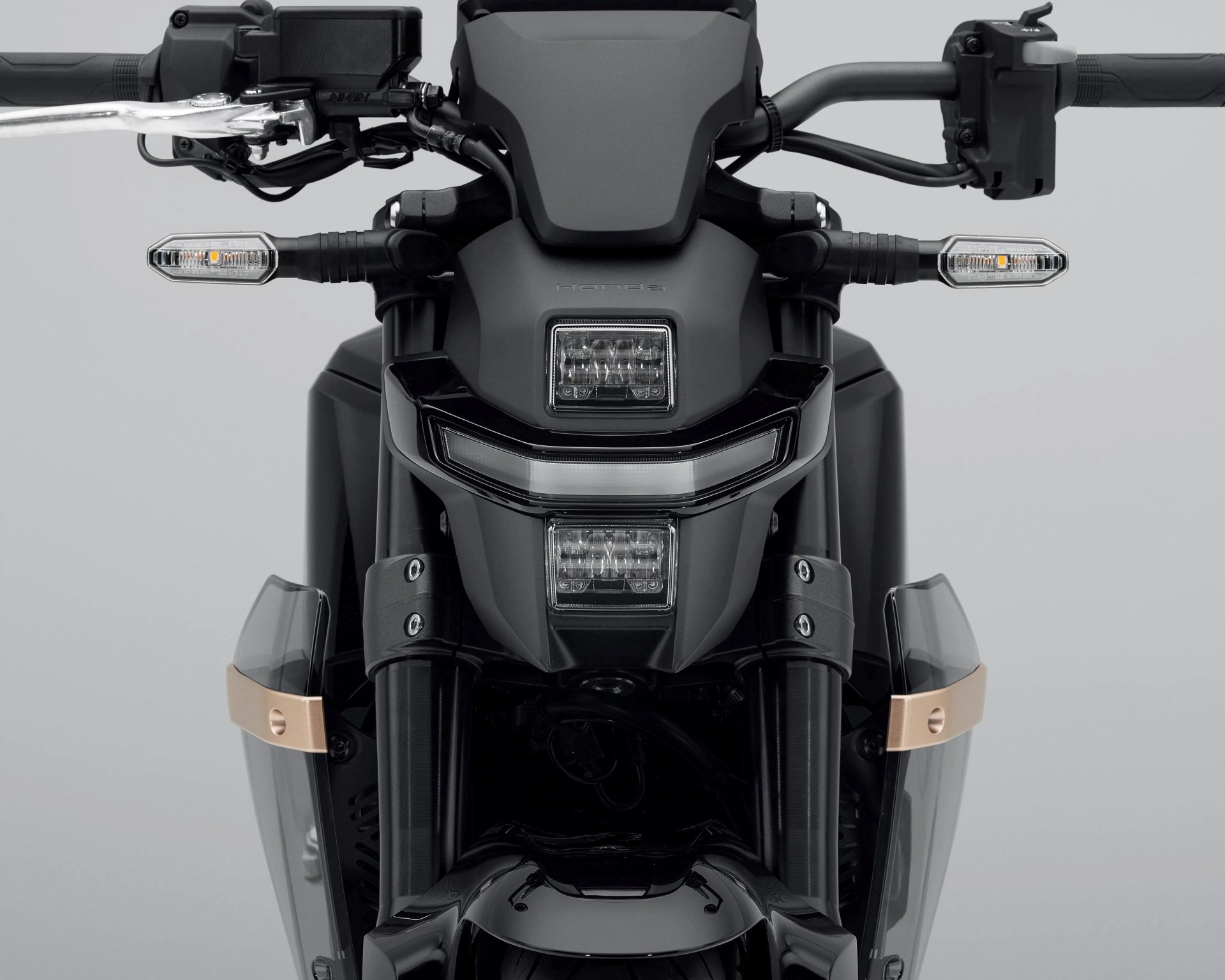 Front view of a black motorcycle featuring a modern, angular headlight design with two rectangular LED lights stacked vertically and a horizontal LED strip in between. The handlebars have clear turn signals with amber bulbs on each side. The front suspension forks are visible, along with small transparent wind deflectors secured by gold-colored brackets on either side. The overall design is sleek and futuristic.