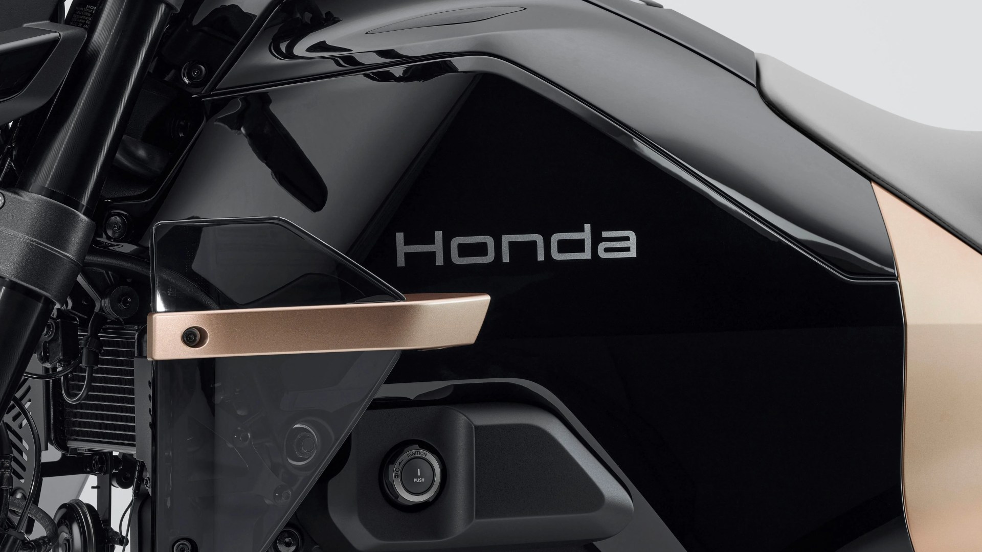 Close-up of a black and gold Honda motorcycle side panel featuring the Honda logo, a transparent wind deflector with a gold mounting bracket, and an ignition push button.