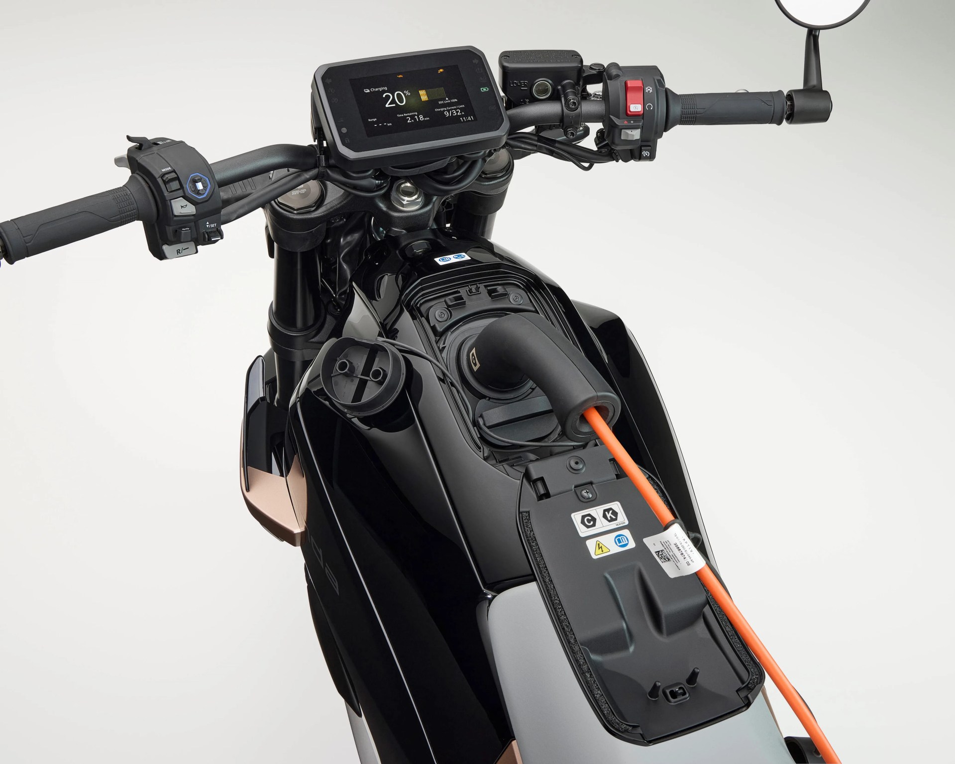 Electric motorcycle dashboard and charging port with an orange charging cable plugged in, showing a digital display indicating 20% charge and other charging details. The motorcycle has black handlebars with control buttons and a rearview mirror on the right side. The charging port is located on the top of the bike's body, beneath a hinged cover.