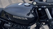Glossy black Honda motorcycle fuel tank with a silver fuel cap and the Honda logo in silver letters on the side. The tank reflects the surrounding environment, including a person taking a photo. Parts of the motorcycle's frame and engine are visible below the tank.