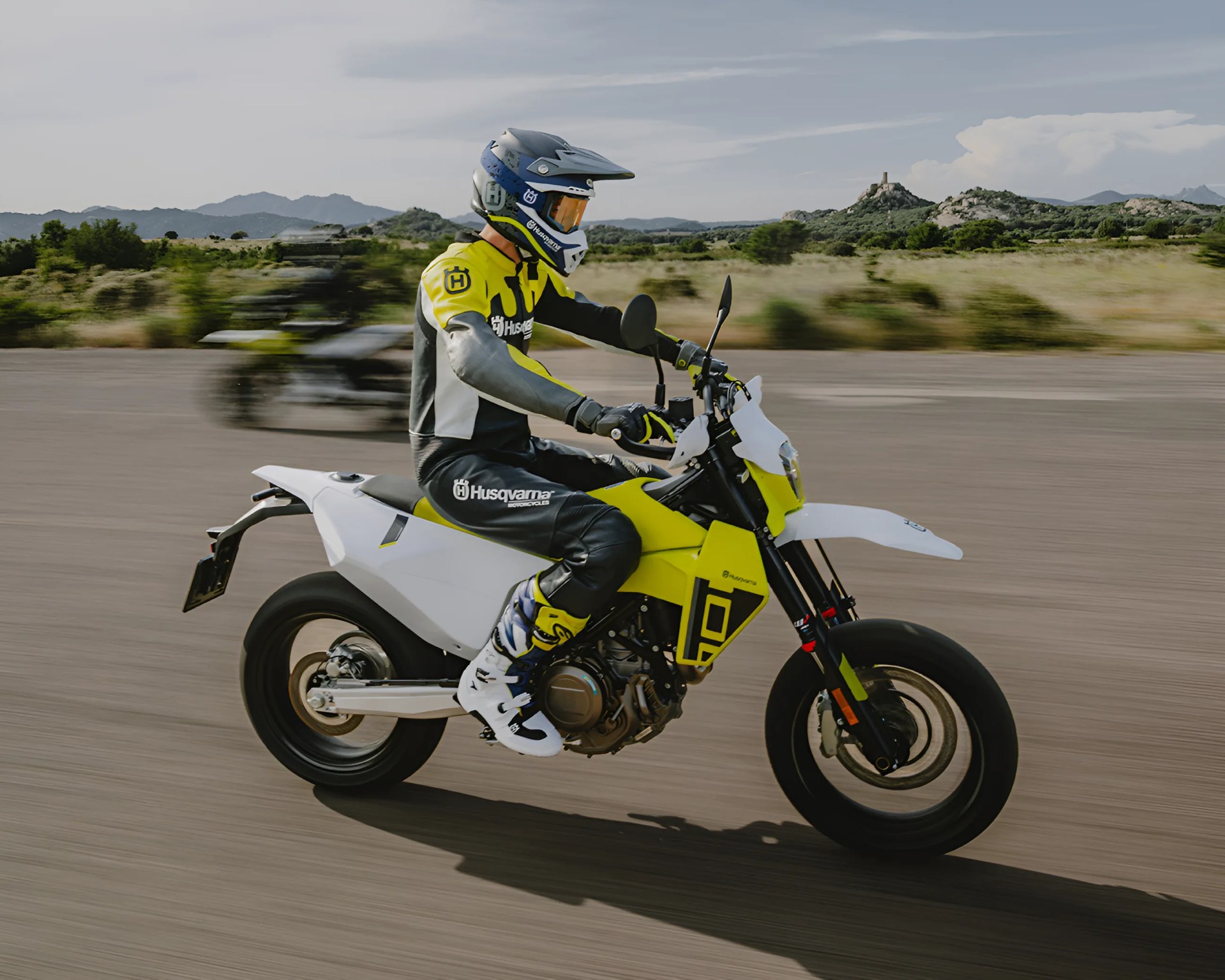 A person wearing a blue and white helmet, yellow and black Husqvarna riding gear, and white boots is riding a yellow and white Husqvarna motorcycle on a paved road with a blurred background of greenery and distant hills.