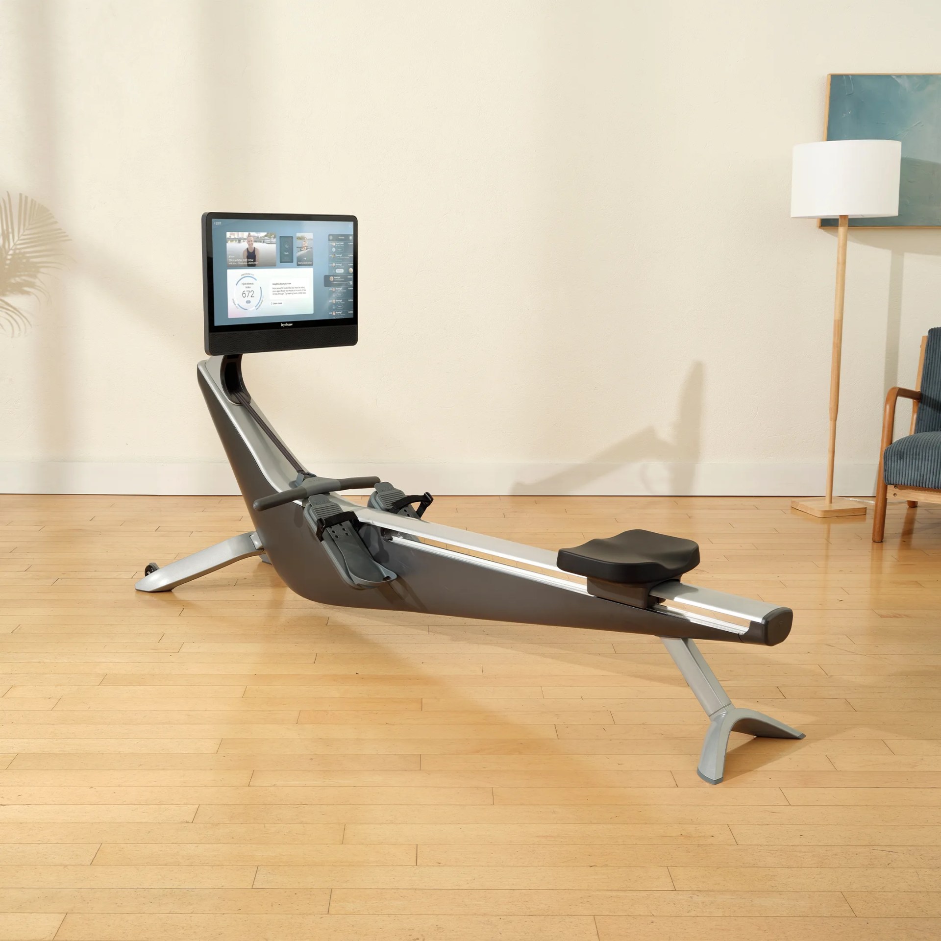 A modern rowing machine with a sleek, gray and silver frame, featuring a black seat and footrests. It has a large touchscreen display mounted at the front, showing workout data and a video interface. The machine is placed on a light wooden floor in a room with a beige wall, a floor lamp, a blue chair, and a piece of artwork partially visible on the wall.