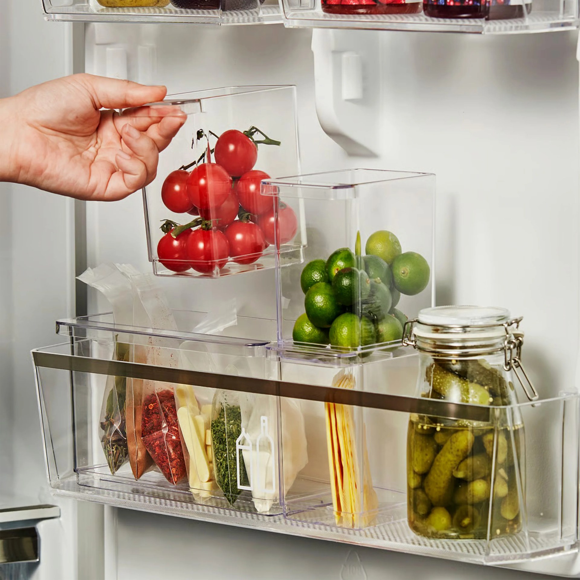 A refrigerator door shelf organized with clear plastic storage containers holding various items. One container is being lifted by a hand and contains red cherry tomatoes. Another container holds small green limes. The shelf also includes a jar of pickles and several small packets of spices or herbs. The containers are neatly arranged to maximize space and visibility.