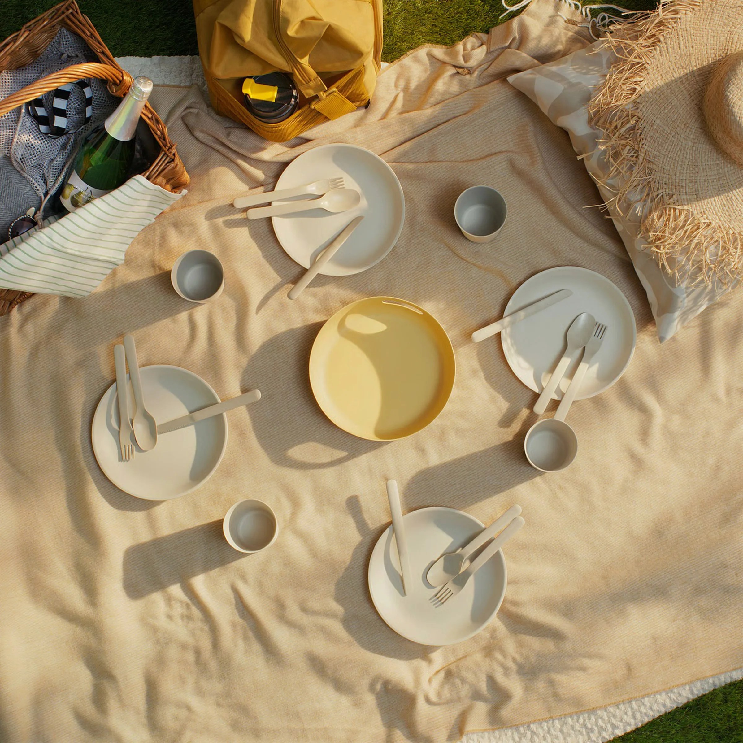 A beige picnic blanket spread on grass with four place settings, each including a beige plate, a knife, fork, and spoon, and a small cup. In the center is a yellow plate. To the left, there is a wicker picnic basket containing a bottle of sparkling beverage and a striped cloth. A mustard yellow bag with a black and yellow water bottle is placed near the top. On the right side, there is a straw hat resting on a beige pillow.