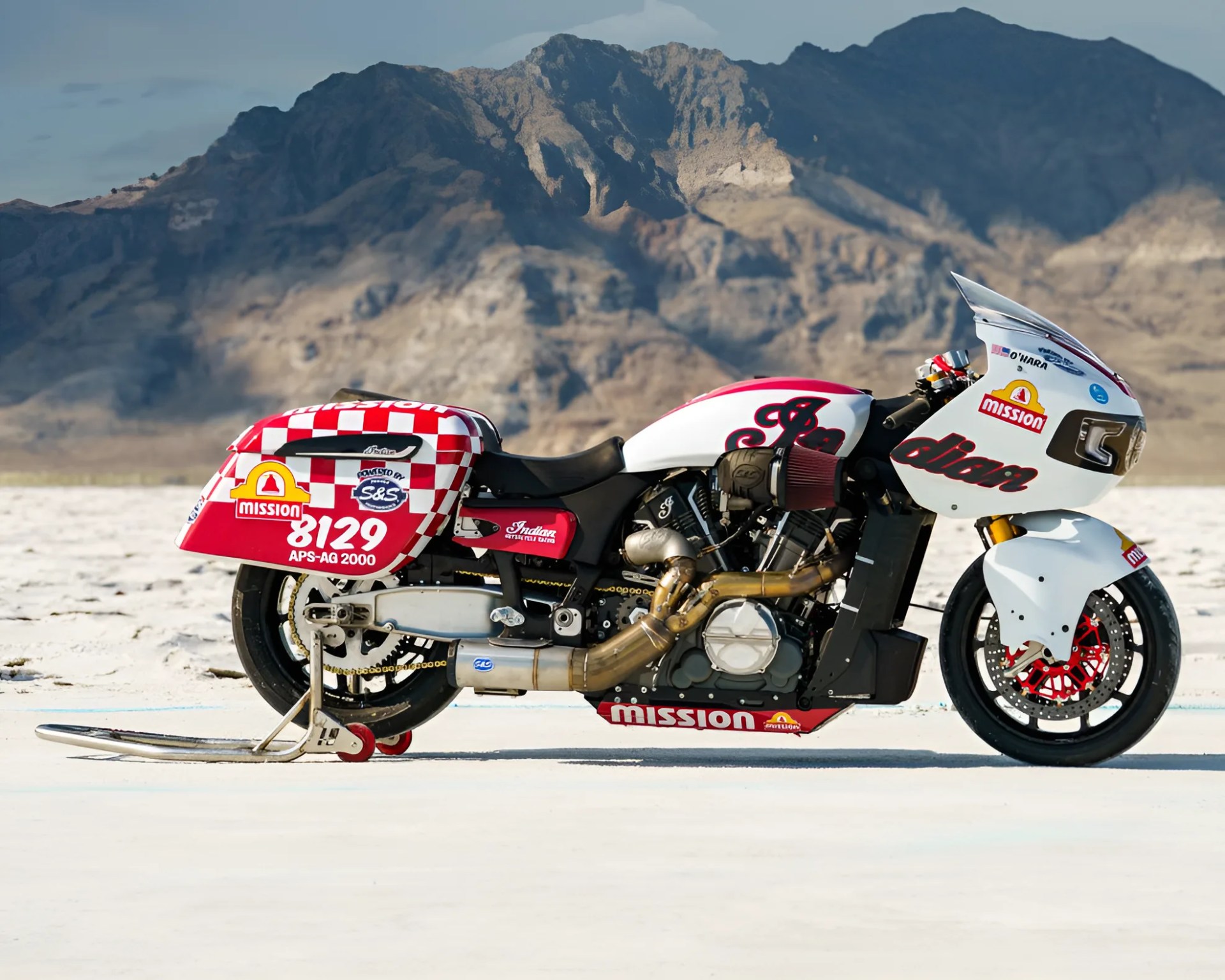 White and red Indian motorcycle with racing decals and exhaust pipes, parked on a salt flat with mountains in the background.