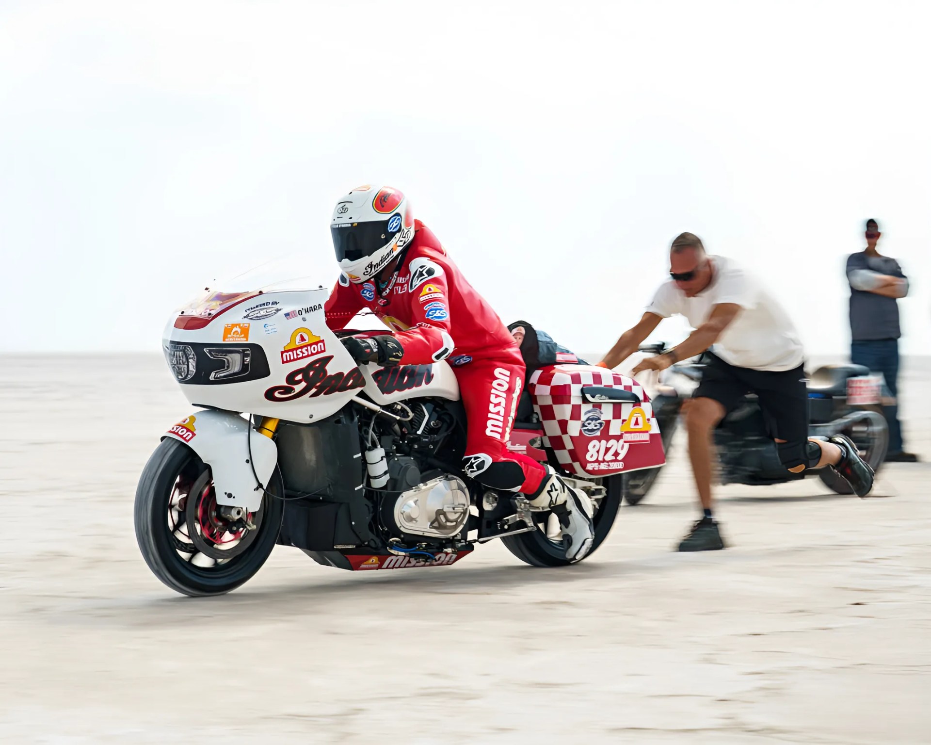 Rider in red racing suit on white and red Indian motorcycle with person pushing from behind on a flat, open surface.