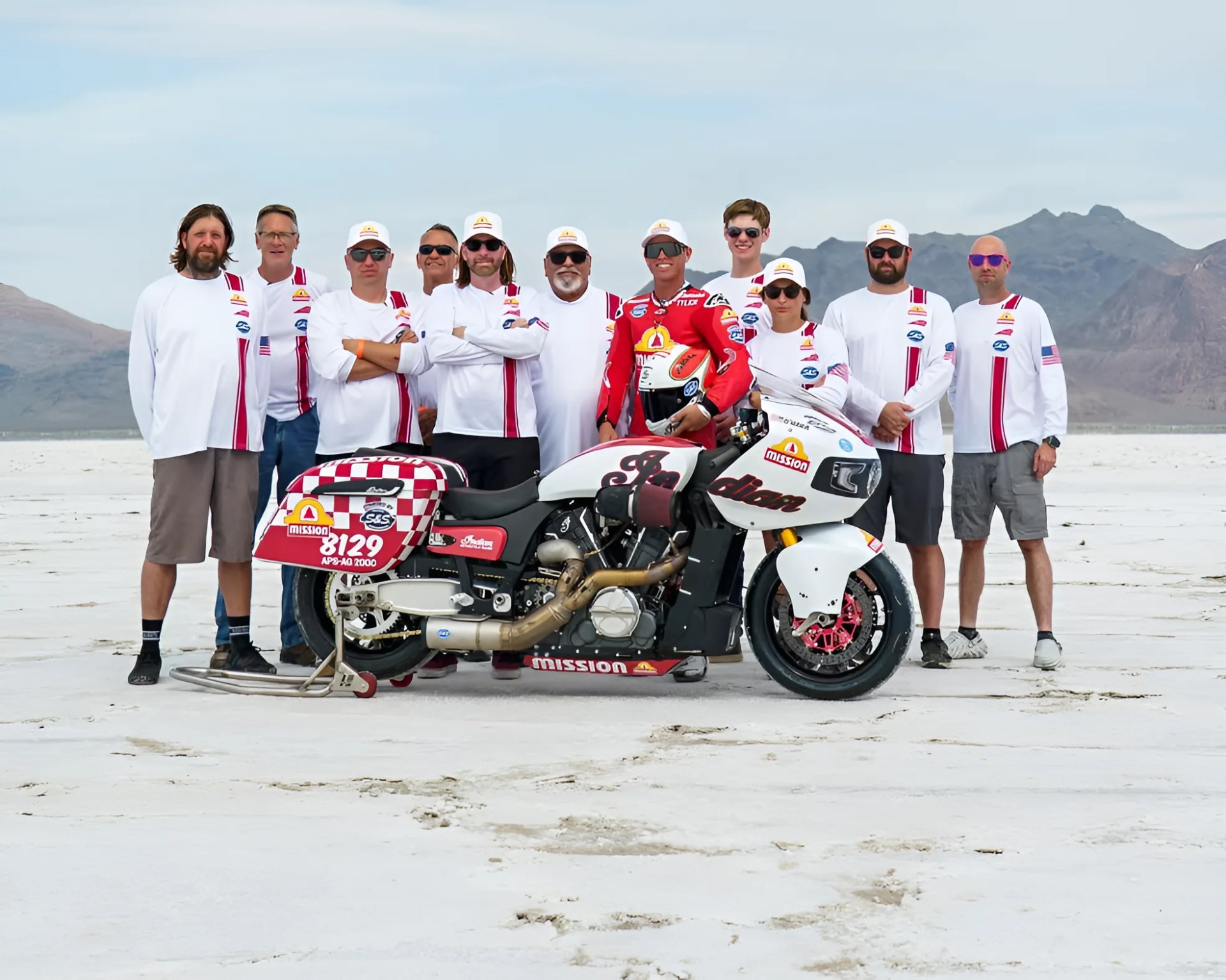 Group of people wearing white shirts and hats with red stripes standing behind a white and red Indian motorcycle on a salt flat with mountains in the background.
