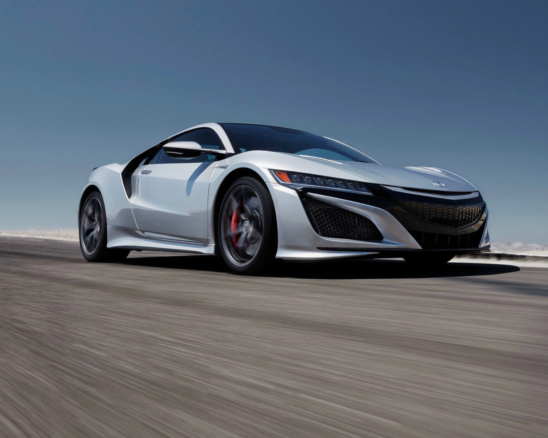 Silver sports car with sleek, aerodynamic design driving on a road, featuring black accents on the front grille and side vents, with red brake calipers visible through the spinning wheels.