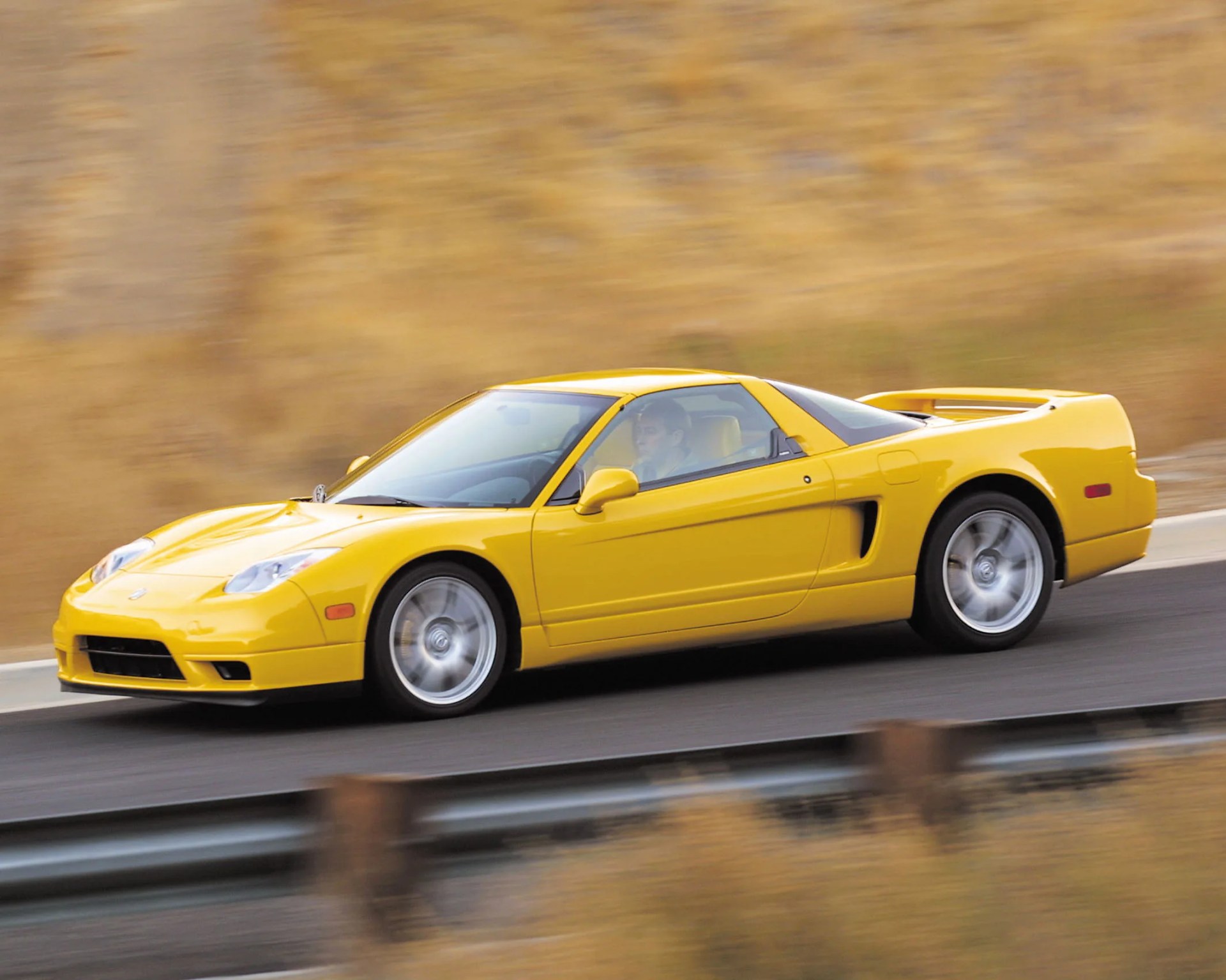Yellow sports car driving on a road with a blurred background, featuring a sleek, low-profile design and silver alloy wheels.