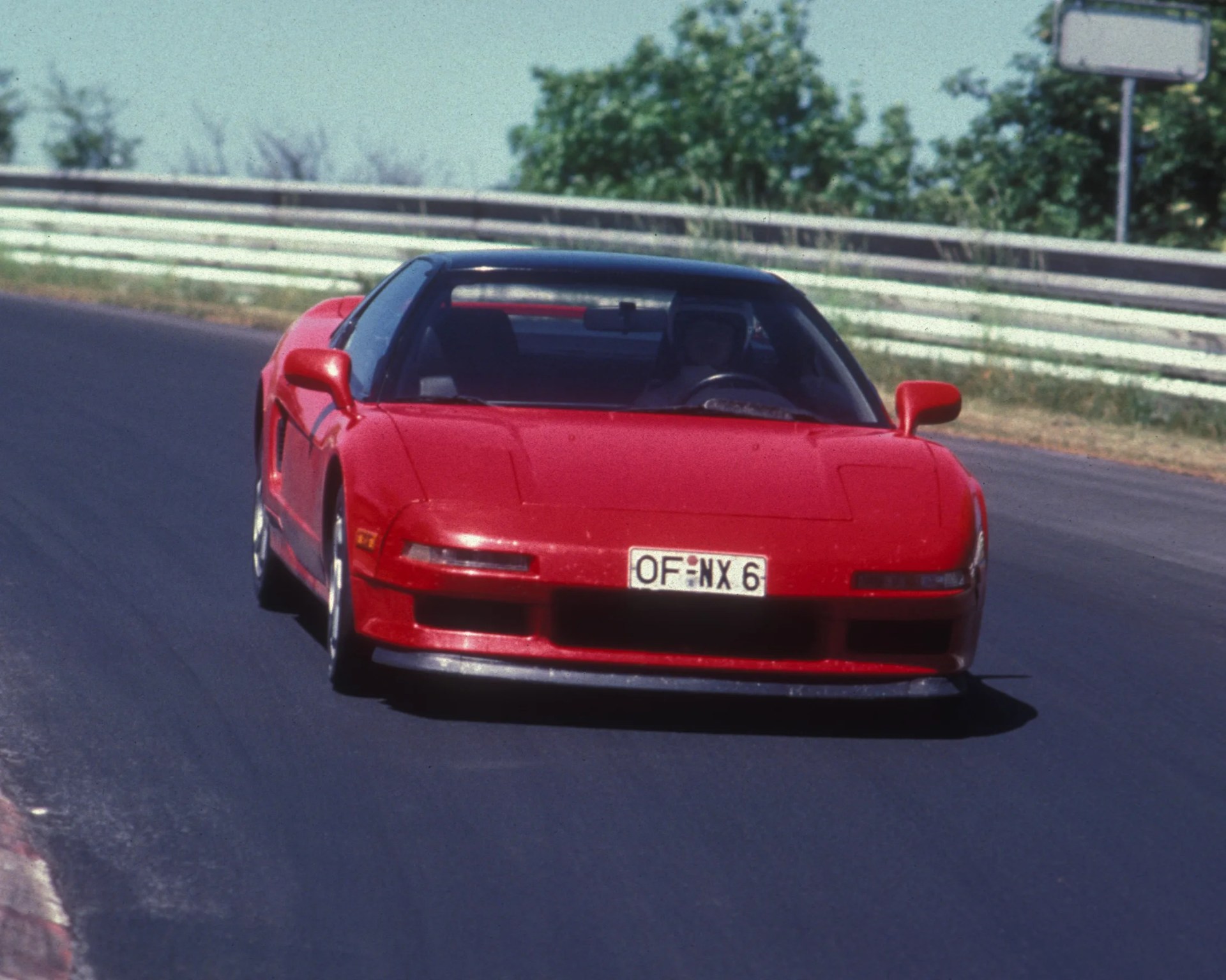 Red sports car with a low front profile driving on a racetrack, featuring pop-up headlights and a visible driver wearing a helmet. The car has a European-style license plate reading "OF NX 6." The background shows a metal guardrail and some greenery.
