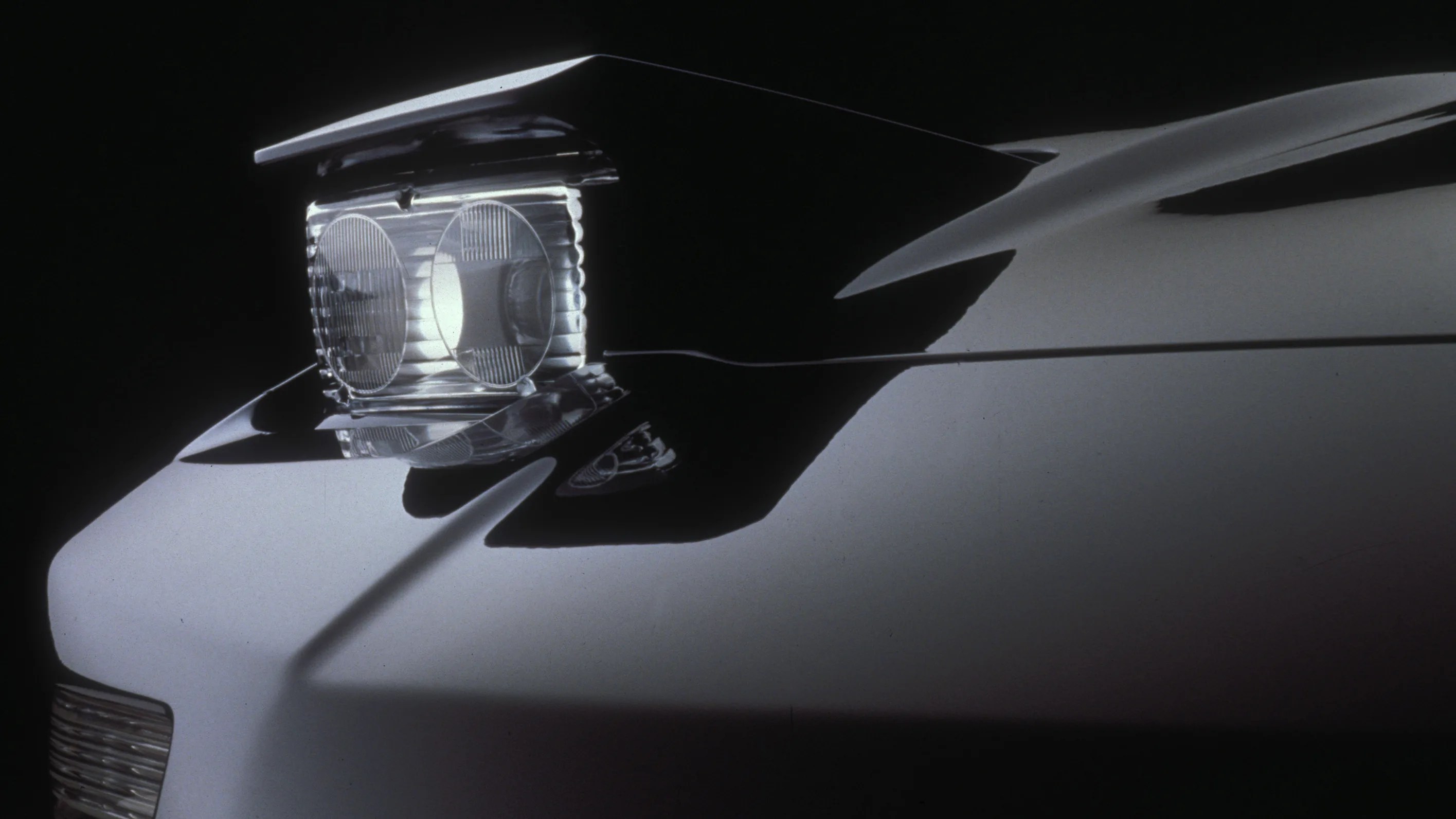 Close-up of a car's front end focusing on a raised, rectangular headlight assembly with two round headlights, set against a dark background. The car's body is smooth and reflective, with a light color.