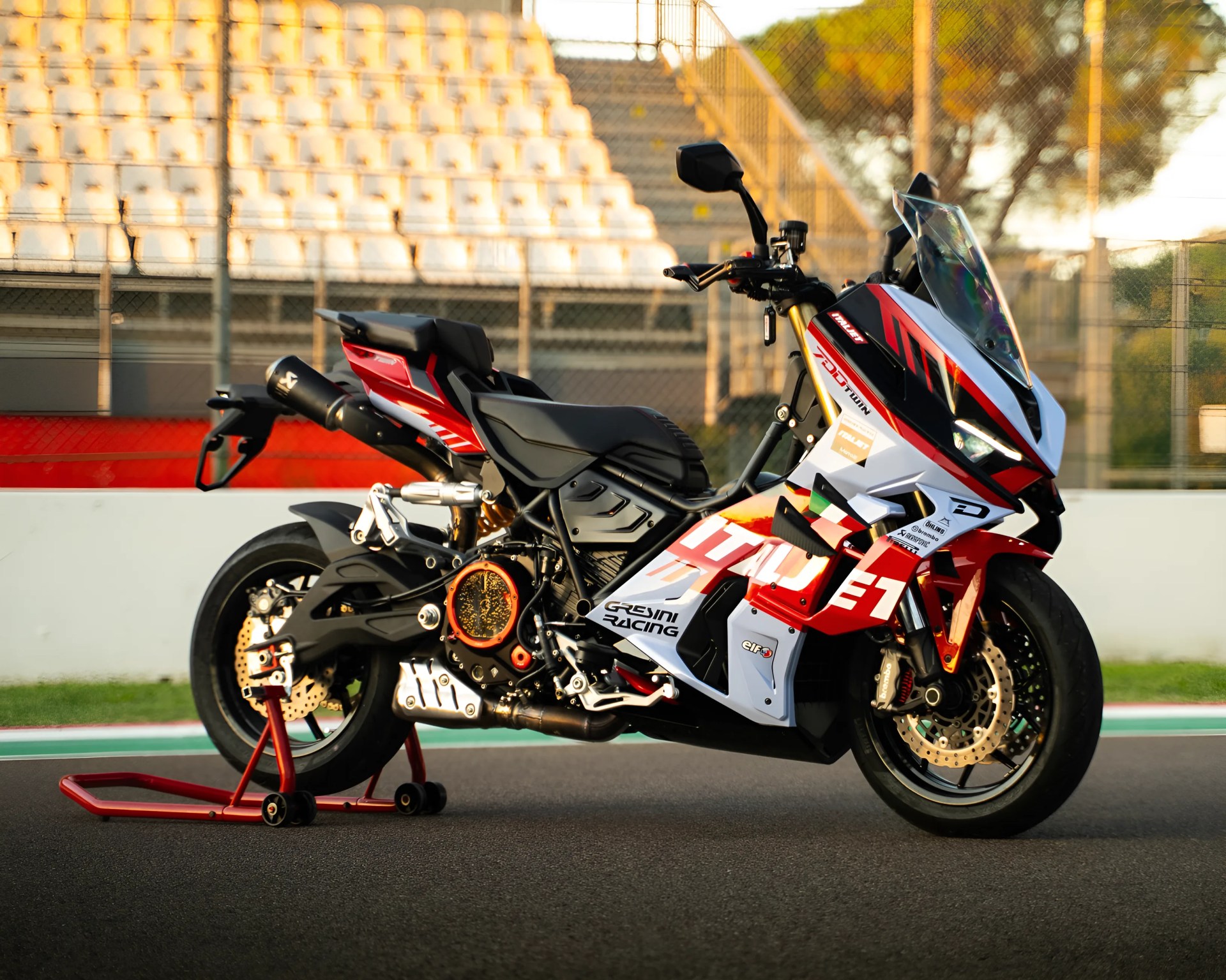 Sport motorcycle with a red, white, and black color scheme featuring racing decals, including "Gresini Racing" and "Italjet." It has a sleek aerodynamic design with a clear windshield, dual front disc brakes, and a black seat. The bike is supported by a red rear stand on a racetrack with empty grandstands in the background.