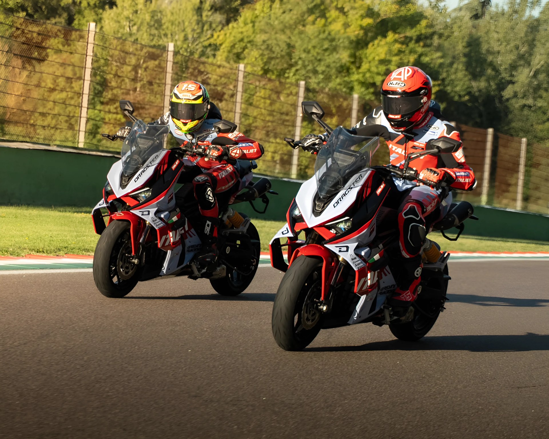 Two motorcyclists in red, white, and black racing suits and helmets are riding sport motorcycles on a paved race track. The motorcycles are primarily white with red and black accents and have the word "DRAGSTER" on the front fairing. The background shows a green barrier and a fence with trees beyond.