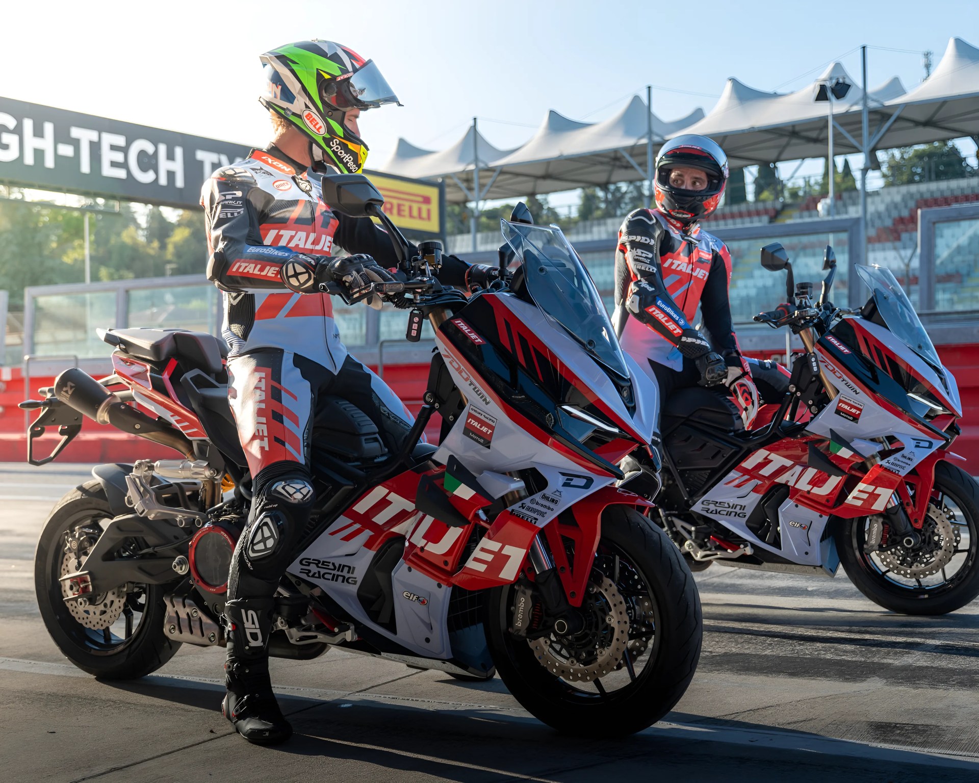 Two motorcyclists in full racing gear and helmets are sitting on red, white, and black Italjet Gresini Racing motorcycles on a racetrack pit lane. The motorcycles have large windshields and prominent branding, and the riders appear to be preparing for a race or practice session. The background shows a grandstand and a sign with "HIGH-TECH" partially visible.