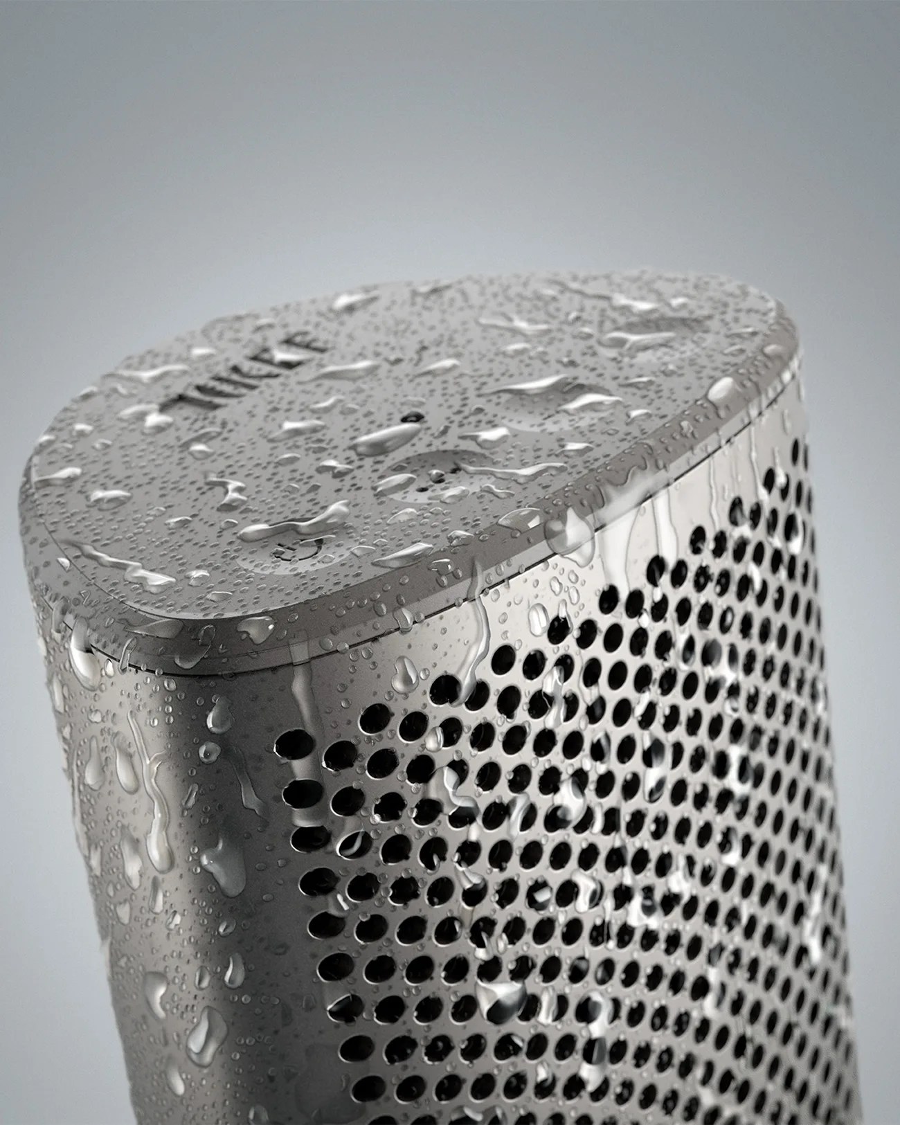 A close-up of a gray, triangular-shaped speaker covered in water droplets, highlighting its perforated grille and smooth top surface.