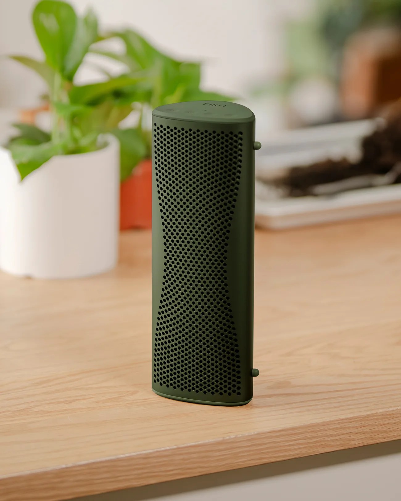Tall, slim, dark green portable speaker with a perforated front grille, standing upright on a light wooden surface. In the background, there are blurred green plants in white and orange pots.