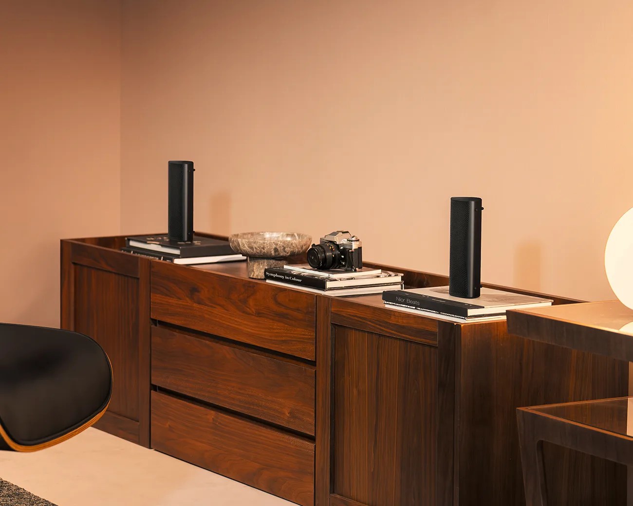 A wooden sideboard with a rich, dark finish featuring three drawers in the center and cabinets on either side. On top of the sideboard are two black cylindrical speakers placed on stacks of books, a vintage camera on a book stack, and a decorative stone bowl. The background wall is painted in a warm beige tone, and part of a black chair and a lamp with a round white shade are visible.