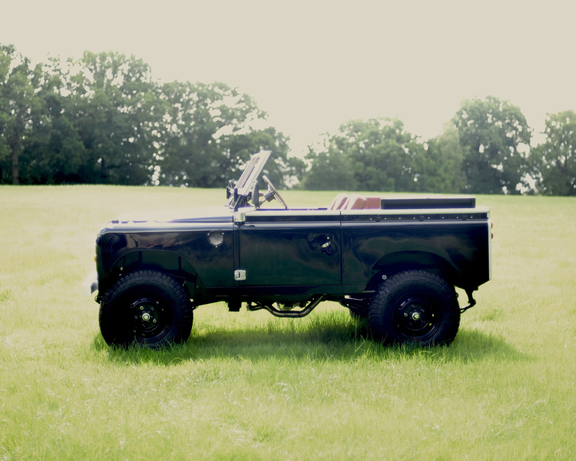 Black vintage off-road vehicle with an open roof and large tires parked on a grassy field, with trees in the background.