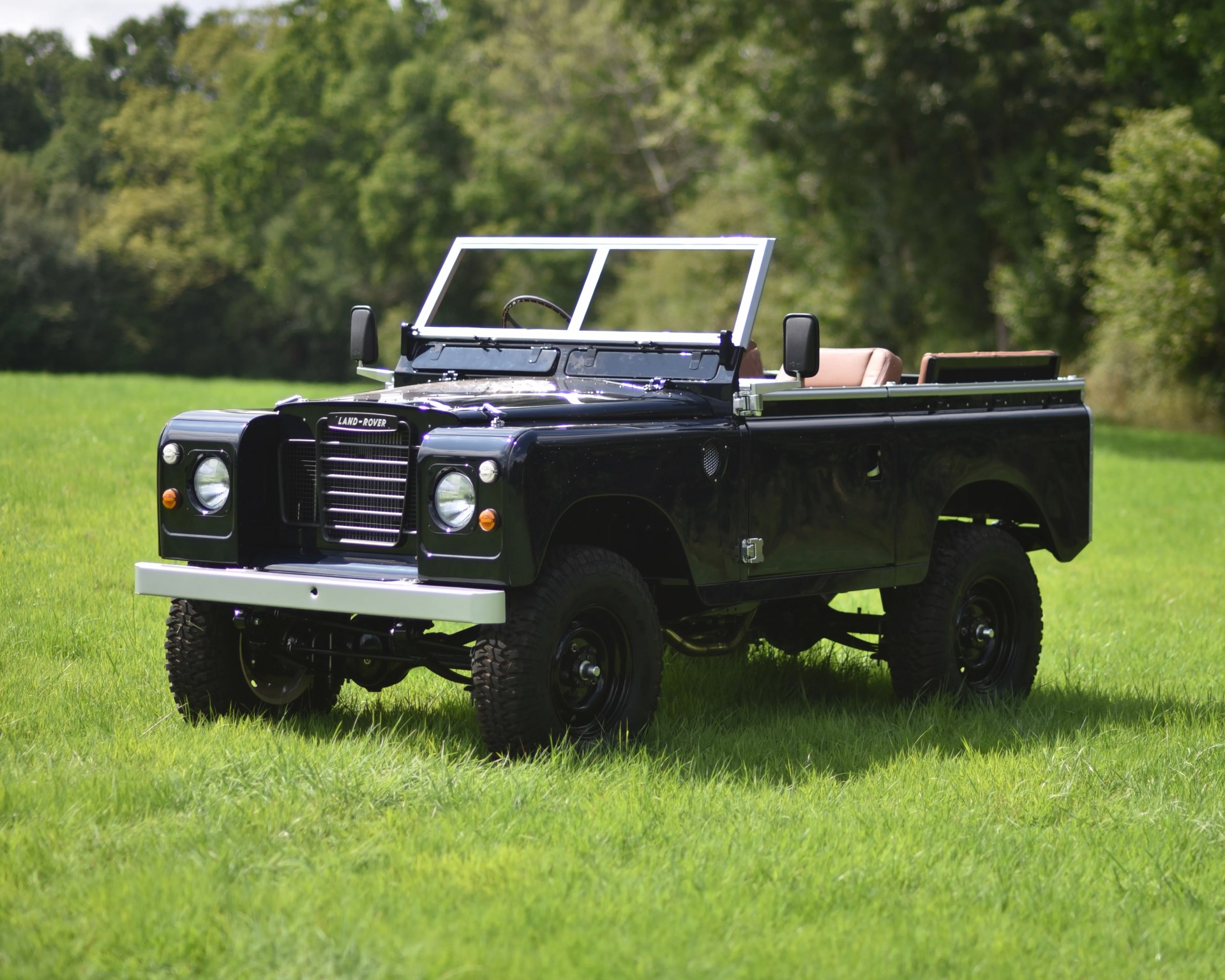 Black classic Land Rover Series vehicle with an open-top design, beige seats, and a silver front bumper, parked on green grass with a background of trees.