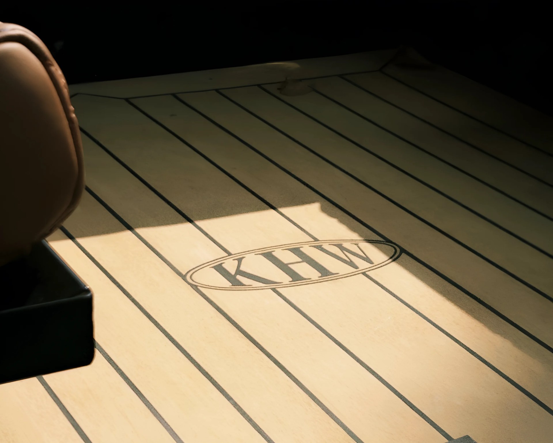A wooden surface with parallel dark lines and an oval logo in the center containing the letters "KHW." The surface is partially shaded, and a brown leather object is visible on the left side.