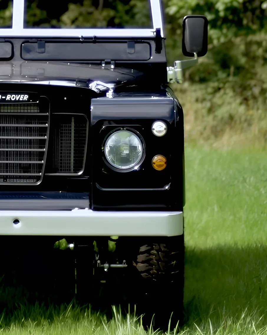 Front view of a black Land Rover vehicle, showing the left headlight, grille, side mirror, and part of the white bumper, parked on green grass with a blurred natural background.