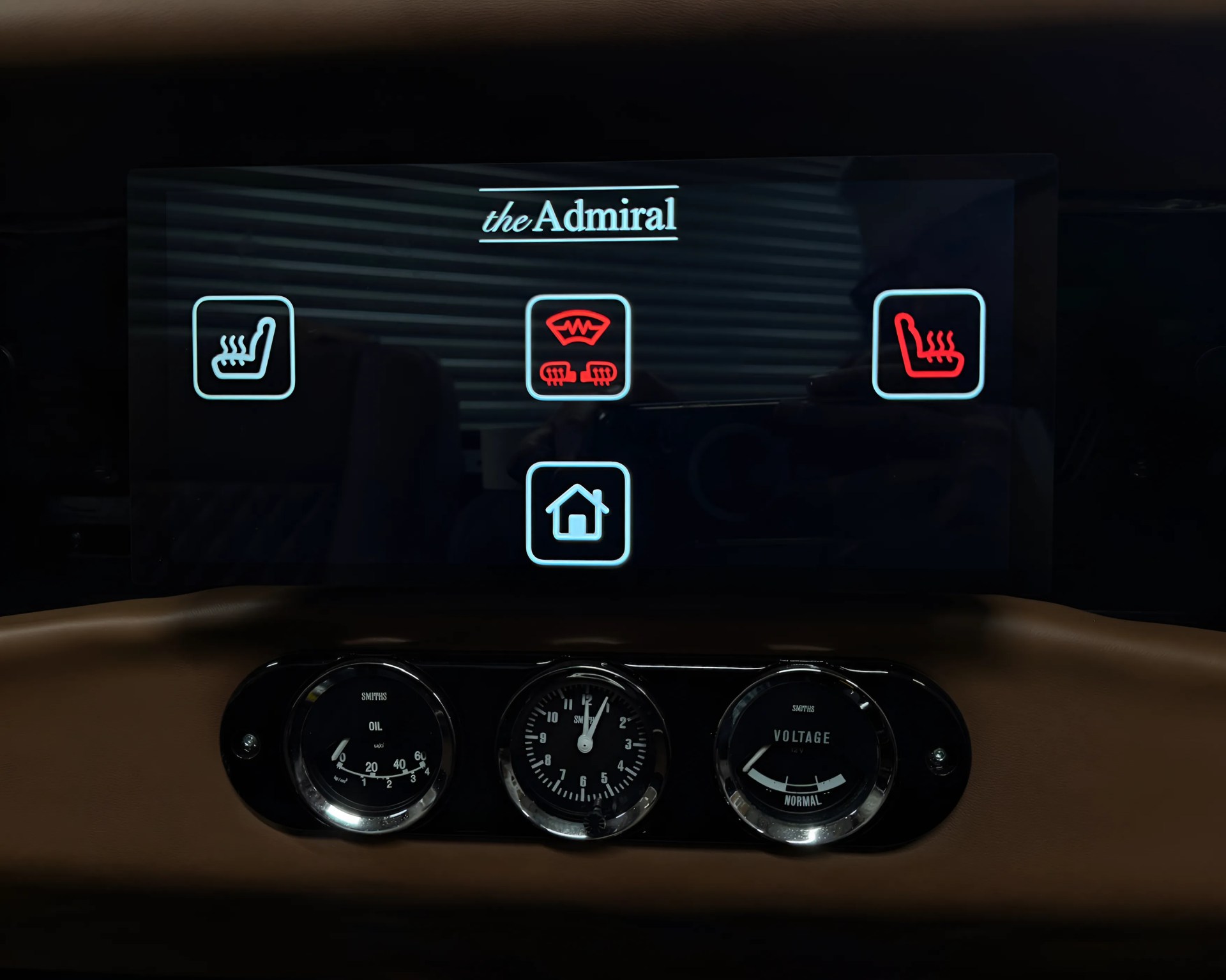 Digital control panel labeled "the Admiral" with icons for heated seats (left and right, right seat icon is red), windshield defrost (center, red), and a home button (bottom center). Below the panel are three analog gauges for oil pressure, a clock, and voltage. The background is dark with a brown dashboard.