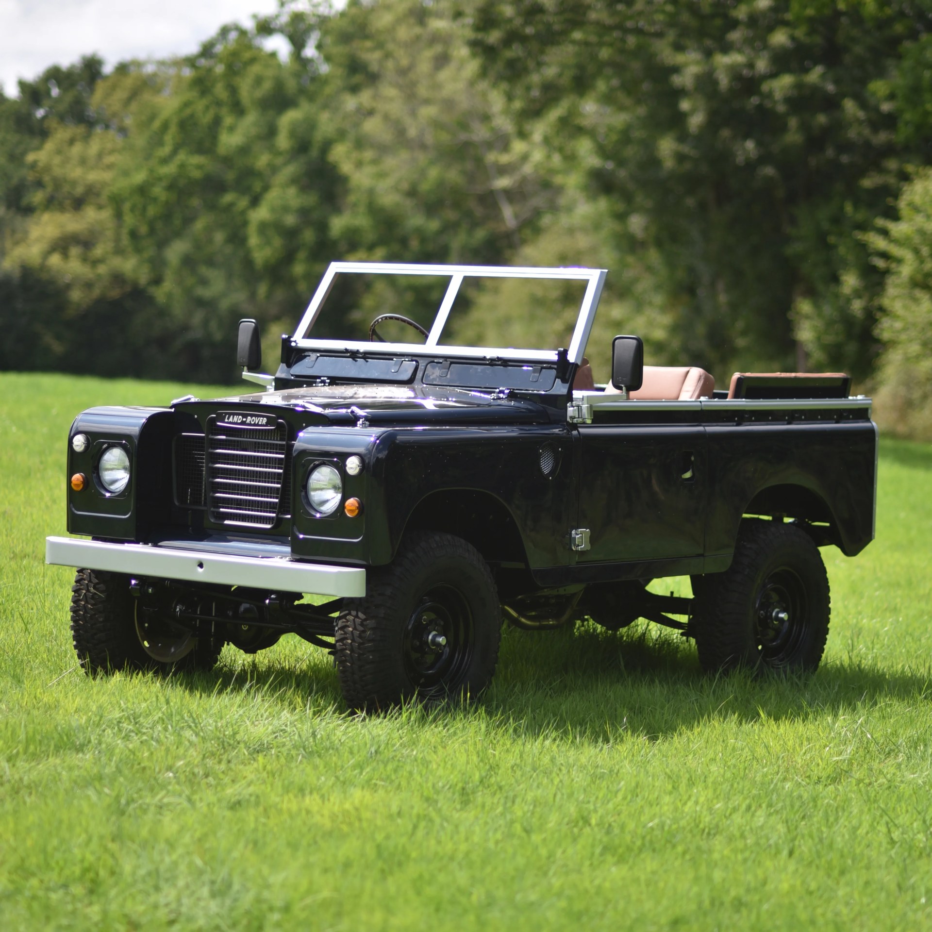 Black vintage Land Rover Series vehicle with an open top, beige seats, and a white front bumper, parked on green grass with trees in the background.