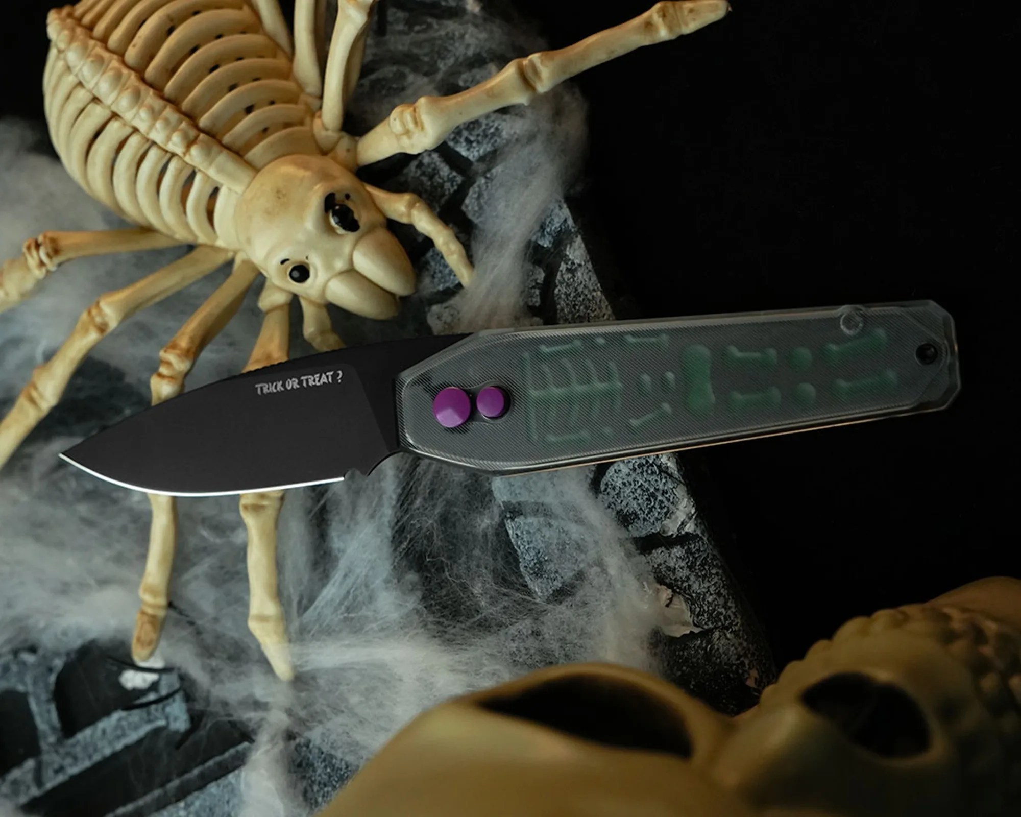 A black folding knife with the phrase "TRICK OR TREAT?" printed on the blade, featuring a translucent handle with visible internal components and two purple buttons. The knife is placed on a surface decorated with fake spider webs, a large skeleton spider, and a skull, creating a Halloween-themed setting.