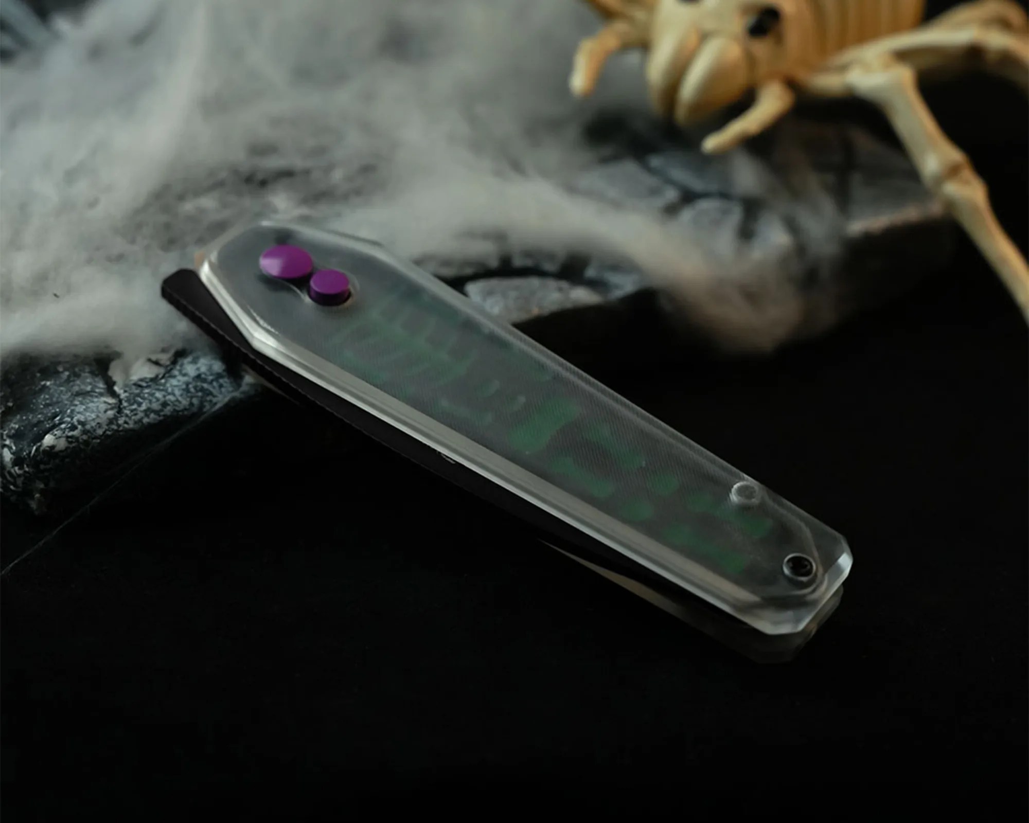 A closed folding knife with a translucent handle featuring a green pattern and two purple buttons, placed on a dark surface with a smoky effect and a partial view of a skeleton decoration in the background.