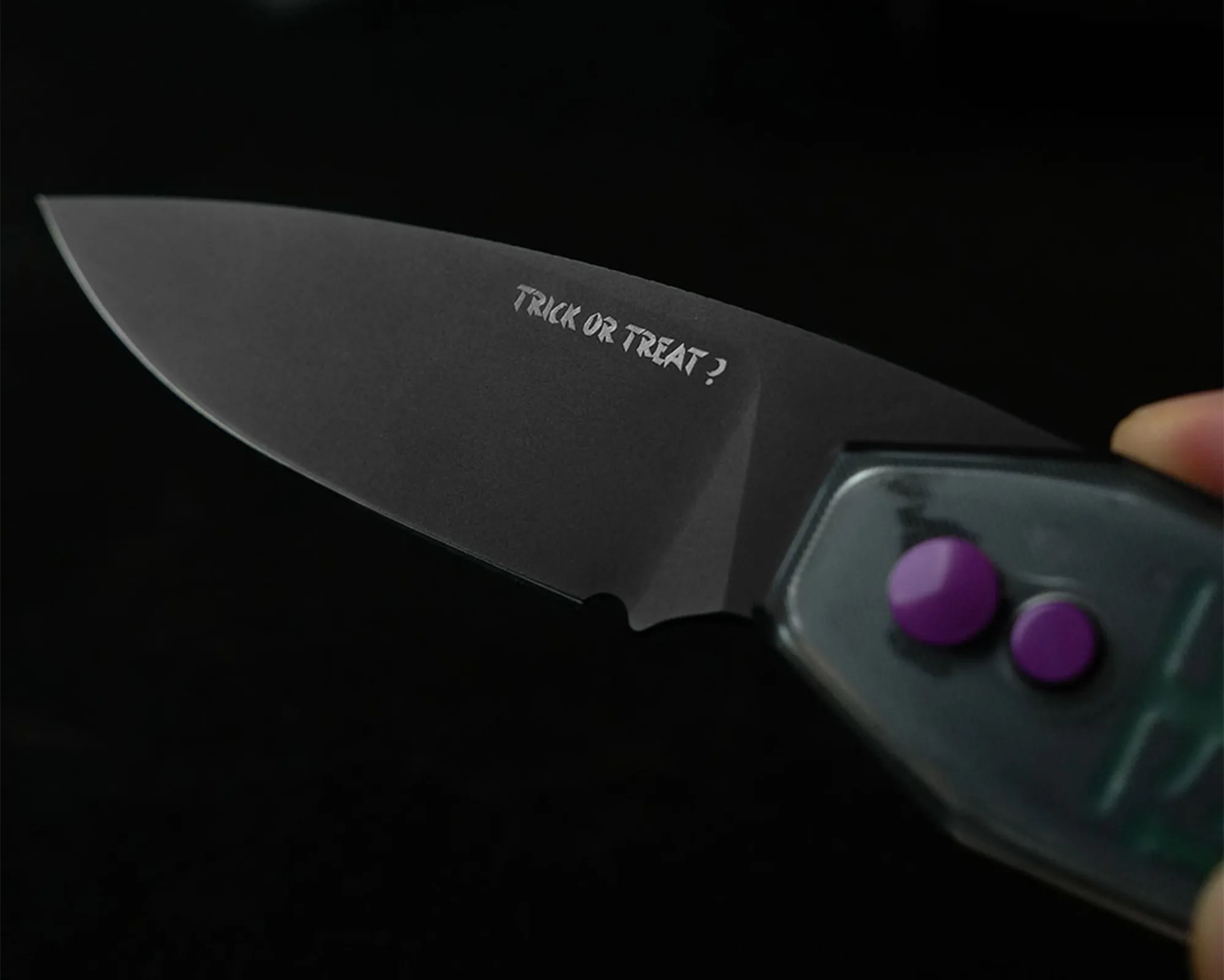 Black folding knife blade with the phrase "TRICK OR TREAT?" etched near the handle, which has two purple buttons.