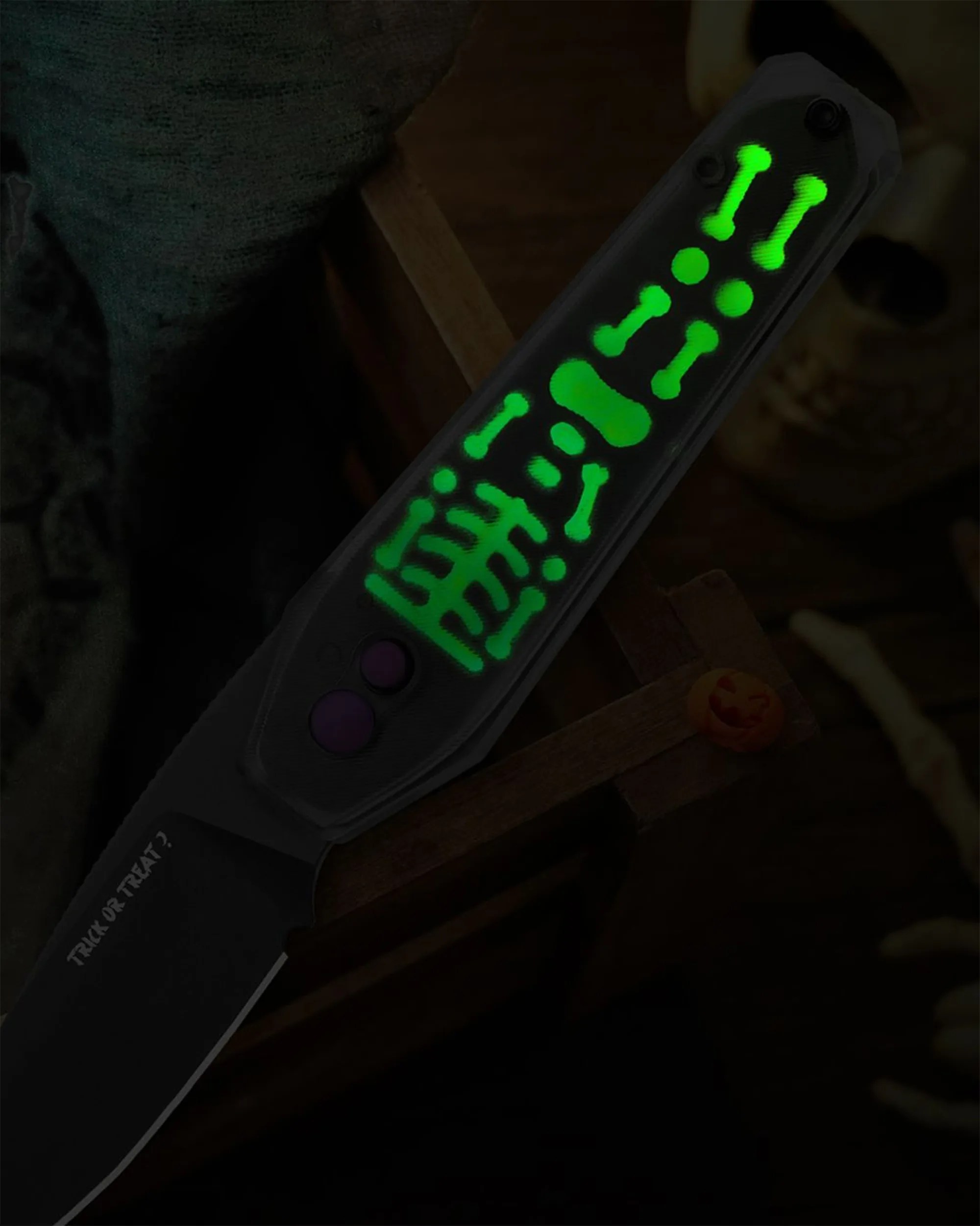 A folding knife with a glowing green skeleton design on the handle. The blade is black with the words "TRICK OR TREAT?" printed near the base. The background includes a wooden surface, a skull, and some bones, creating a spooky Halloween theme.