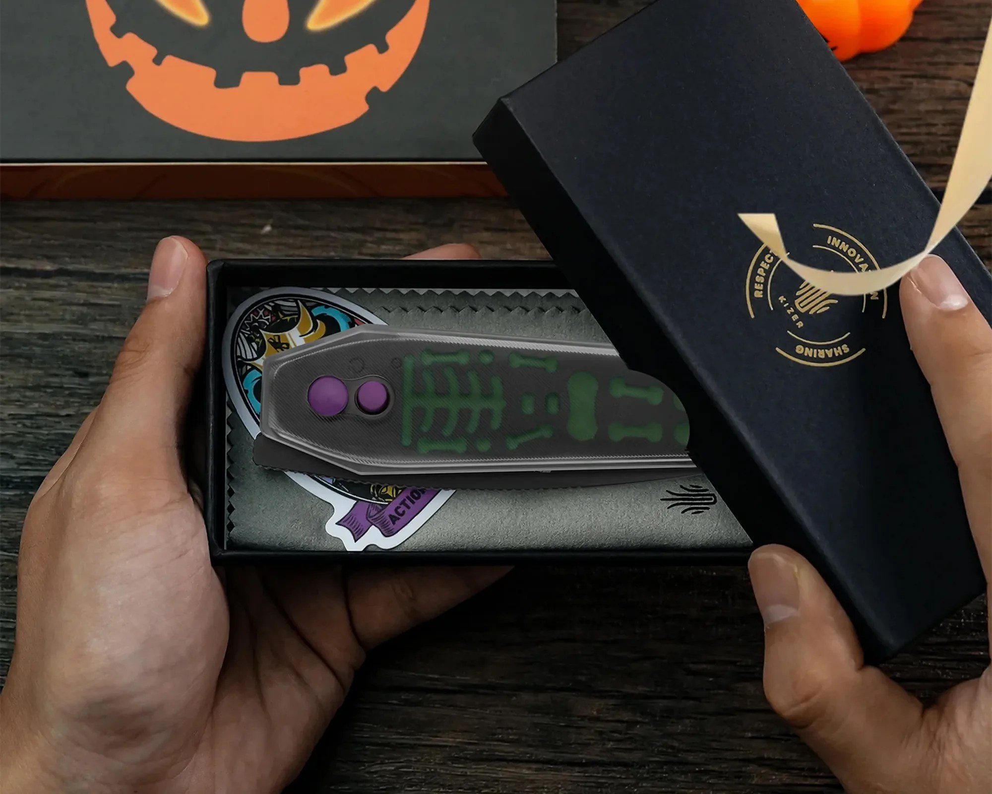A hand holding a black box with a partially opened lid revealing a folding knife inside. The knife has a gray handle with green skeleton-like patterns and two purple buttons. There is also a colorful sticker with a skull design and the word "ACTION" inside the box. The box lid has a gold circular logo with the words "RESPECT," "INNOVATION," and "SHARING" around a handshake symbol. The background includes a wooden surface and part of an orange pumpkin decoration.