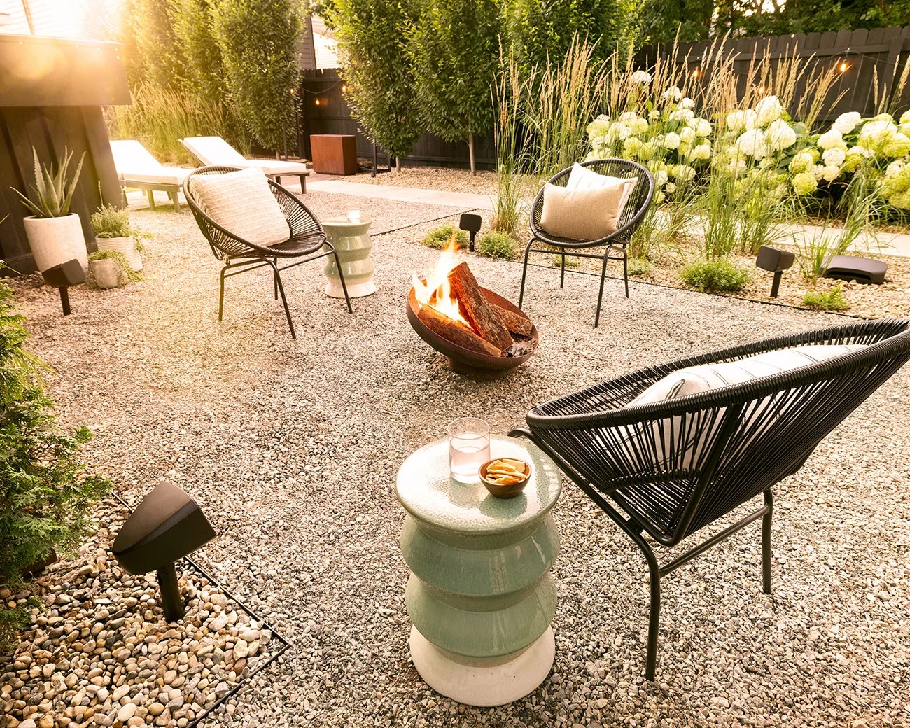 Outdoor seating area with three black woven chairs, each with a light-colored cushion, arranged around a small fire pit with burning logs. The ground is covered with small gravel, and there are two green ceramic side tables, one holding a glass of water and a small bowl of snacks. The background features tall grasses, white hydrangea flowers, and a dark wooden fence, with soft sunlight filtering through the trees.