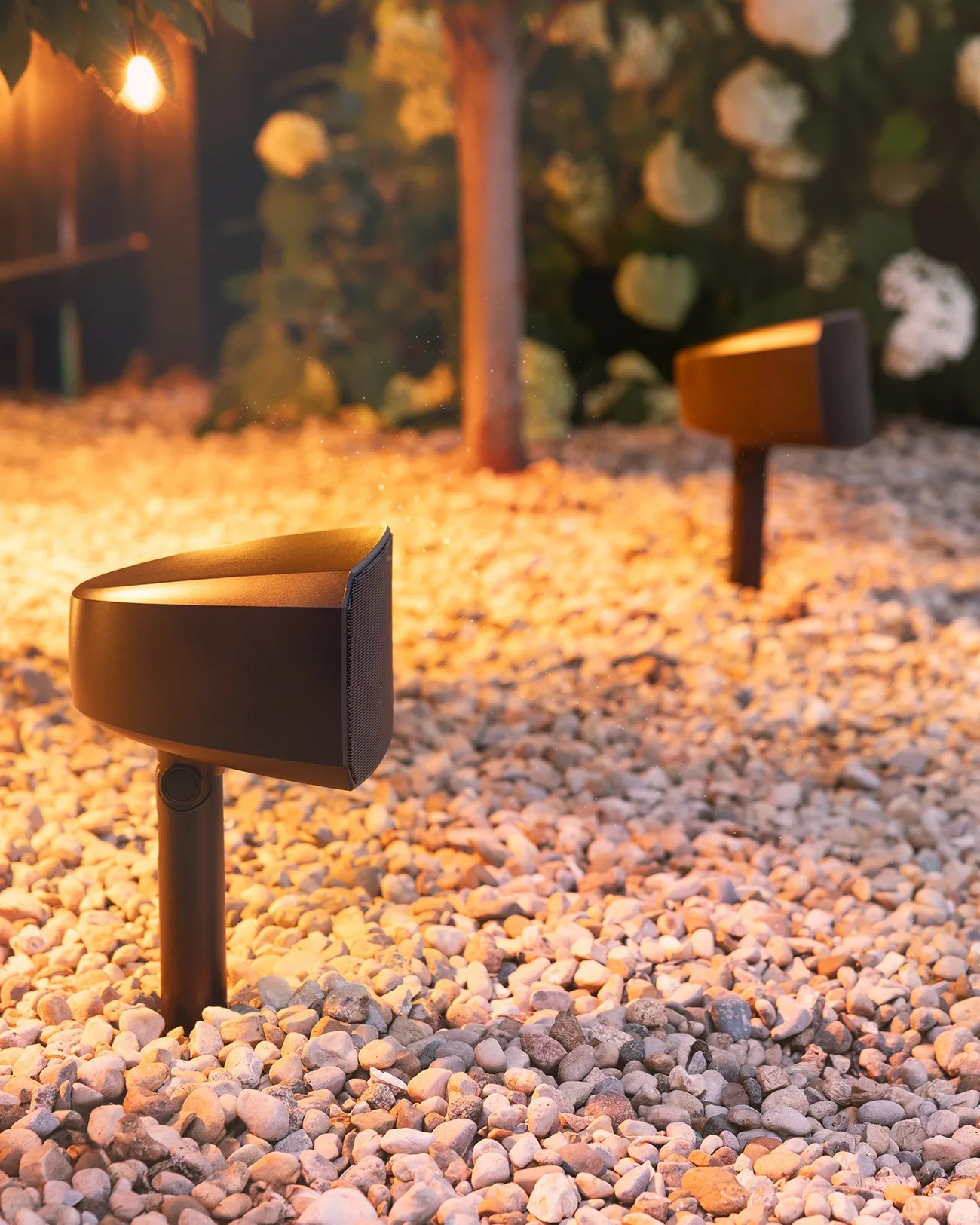 Two modern outdoor speakers with a sleek, black design are installed on stakes in a gravel-covered garden area, illuminated by warm ambient lighting. The background features blurred greenery and white flowers.