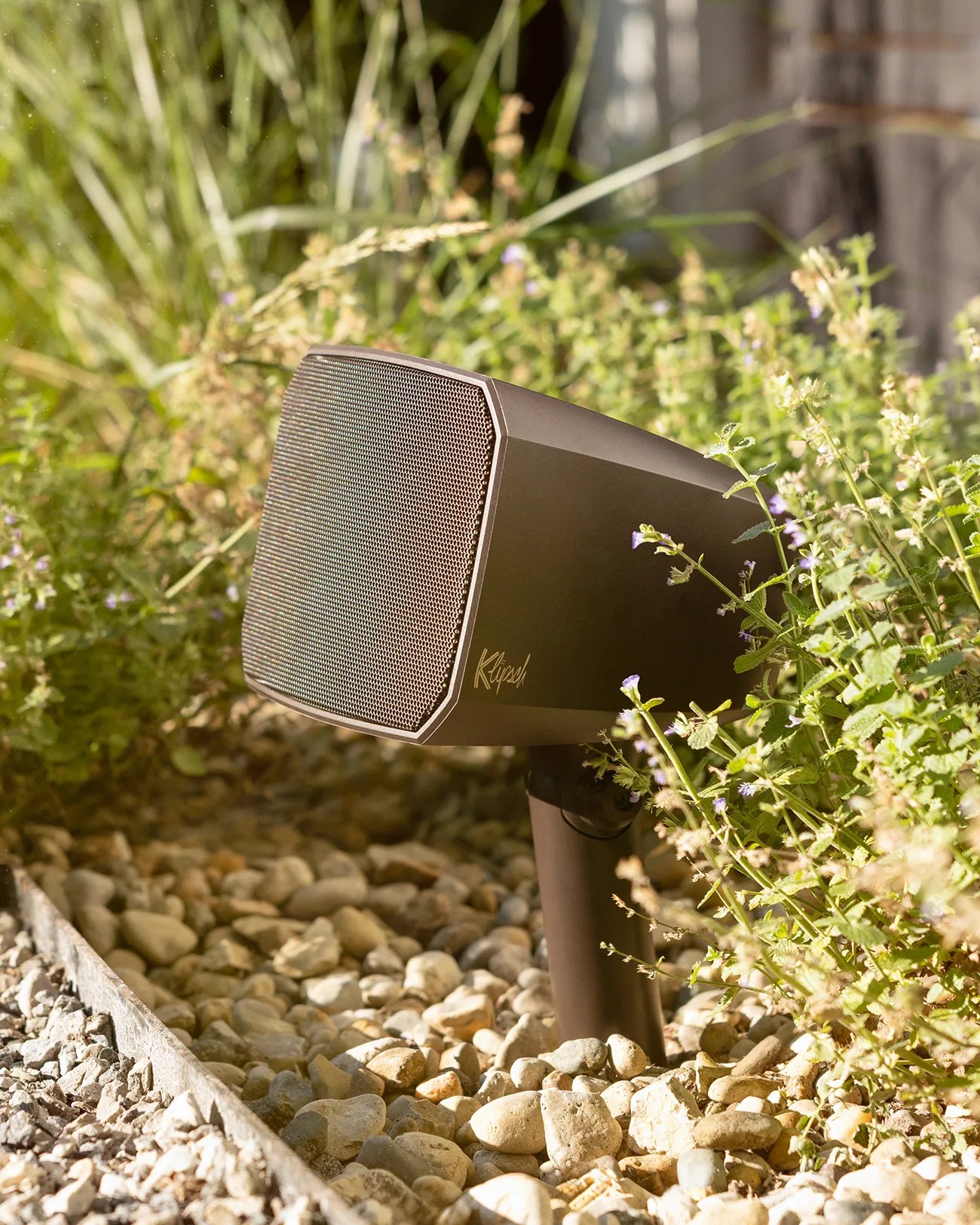 Outdoor black speaker mounted on a stake, positioned among green plants and small rocks, with sunlight illuminating the scene. The speaker has a mesh front and the brand name "Klipsch" visible on the side.